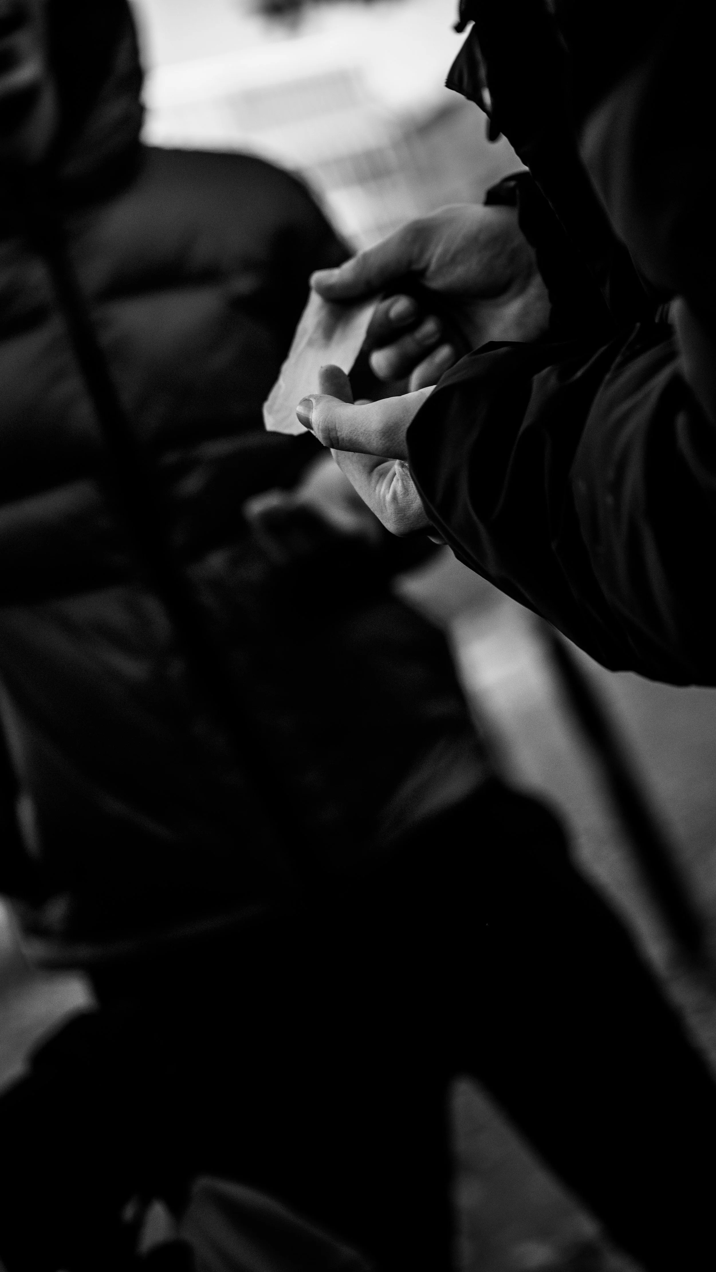 Close-up of a person's hands holding a small paper note, with other people visible in the background, all wrapped in jackets, in black and white.