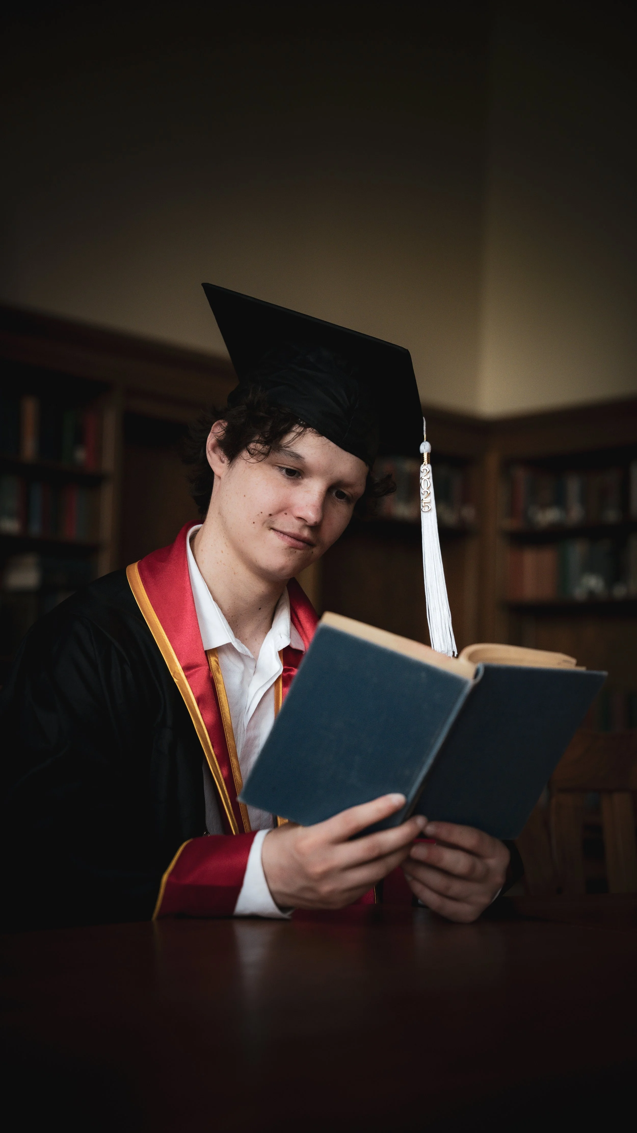 Young man in graduation cap and gown reading a book in a library.