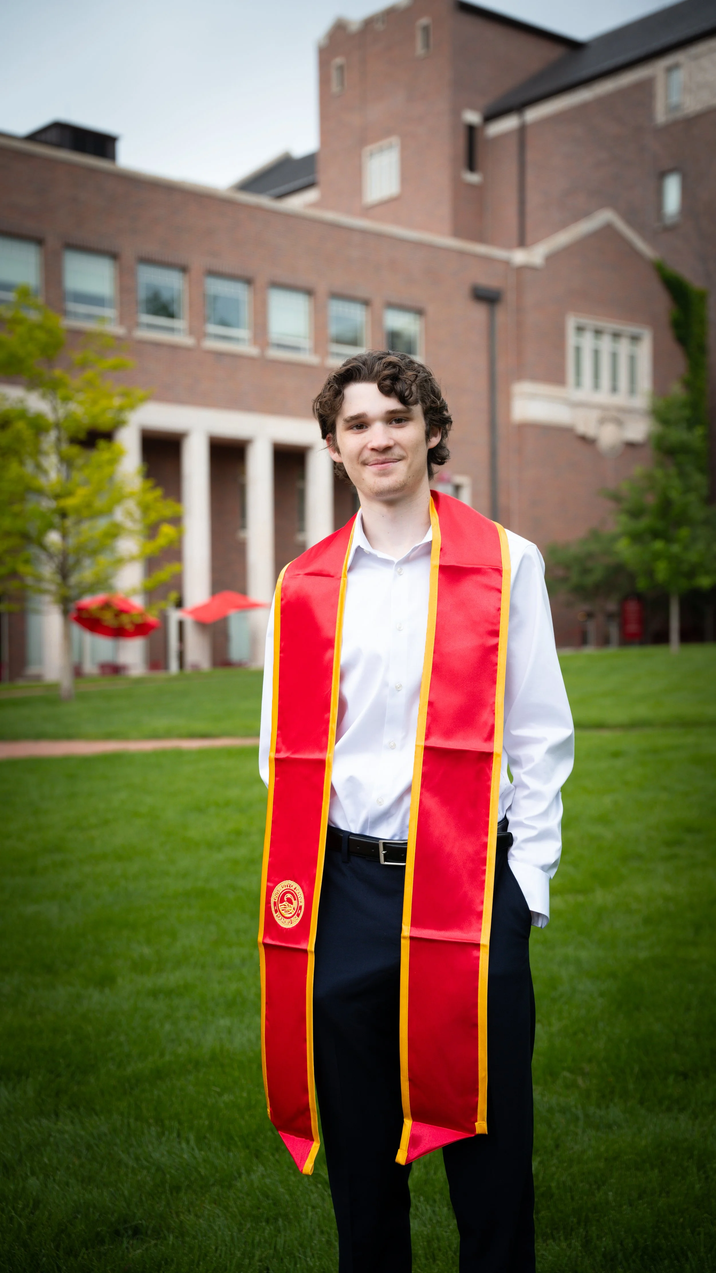 A young man in a white shirt and black pants standing on a grassy lawn outside a brick building, wearing a red graduation stole with yellow edging.