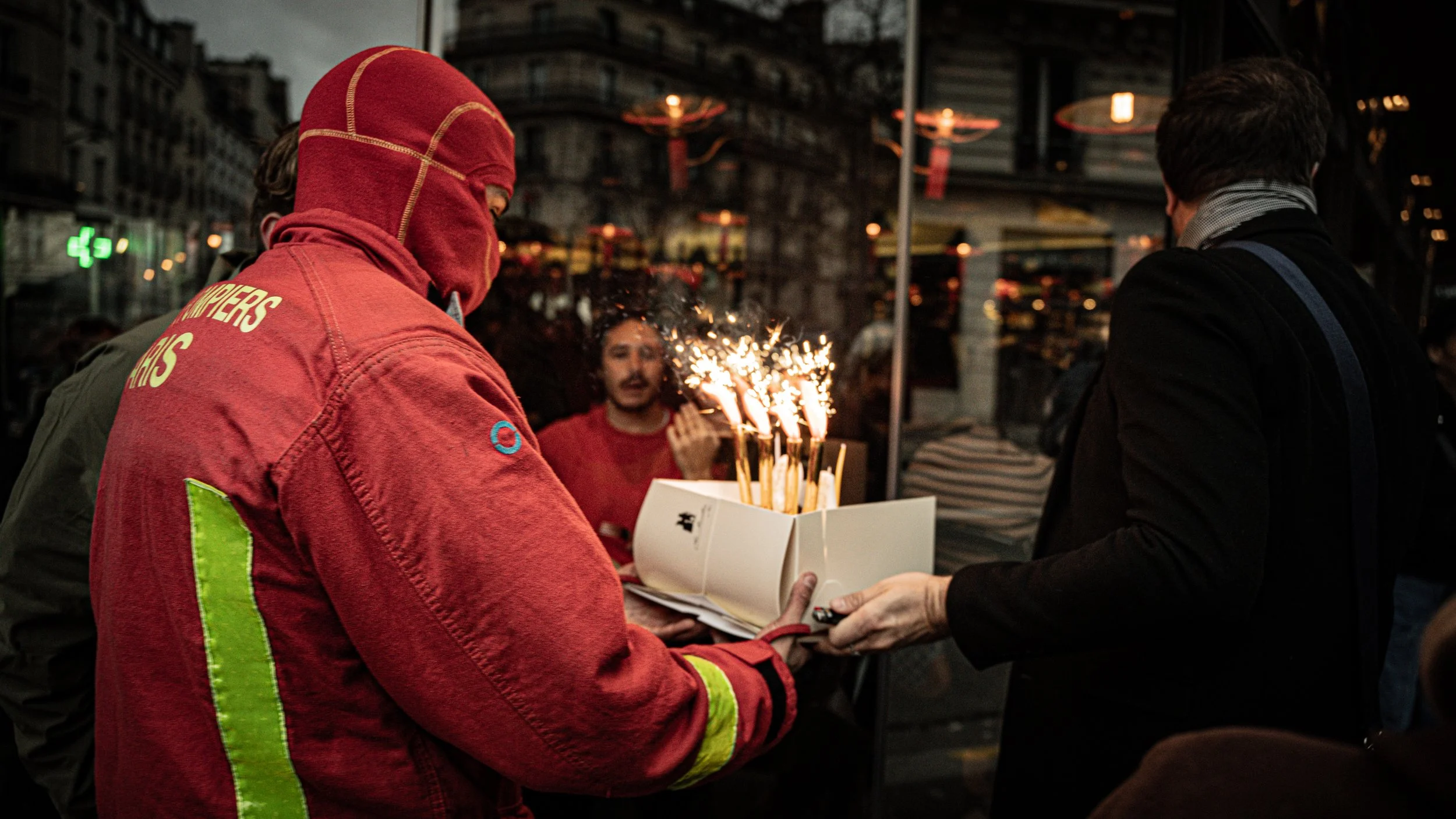 A person in a red uniform with a hood and face covering holding a box with sparklers, standing outside at night. Another person in a dark coat is handing the box to him. In the background, a man with a mustache in a red sweater is looking at the spar