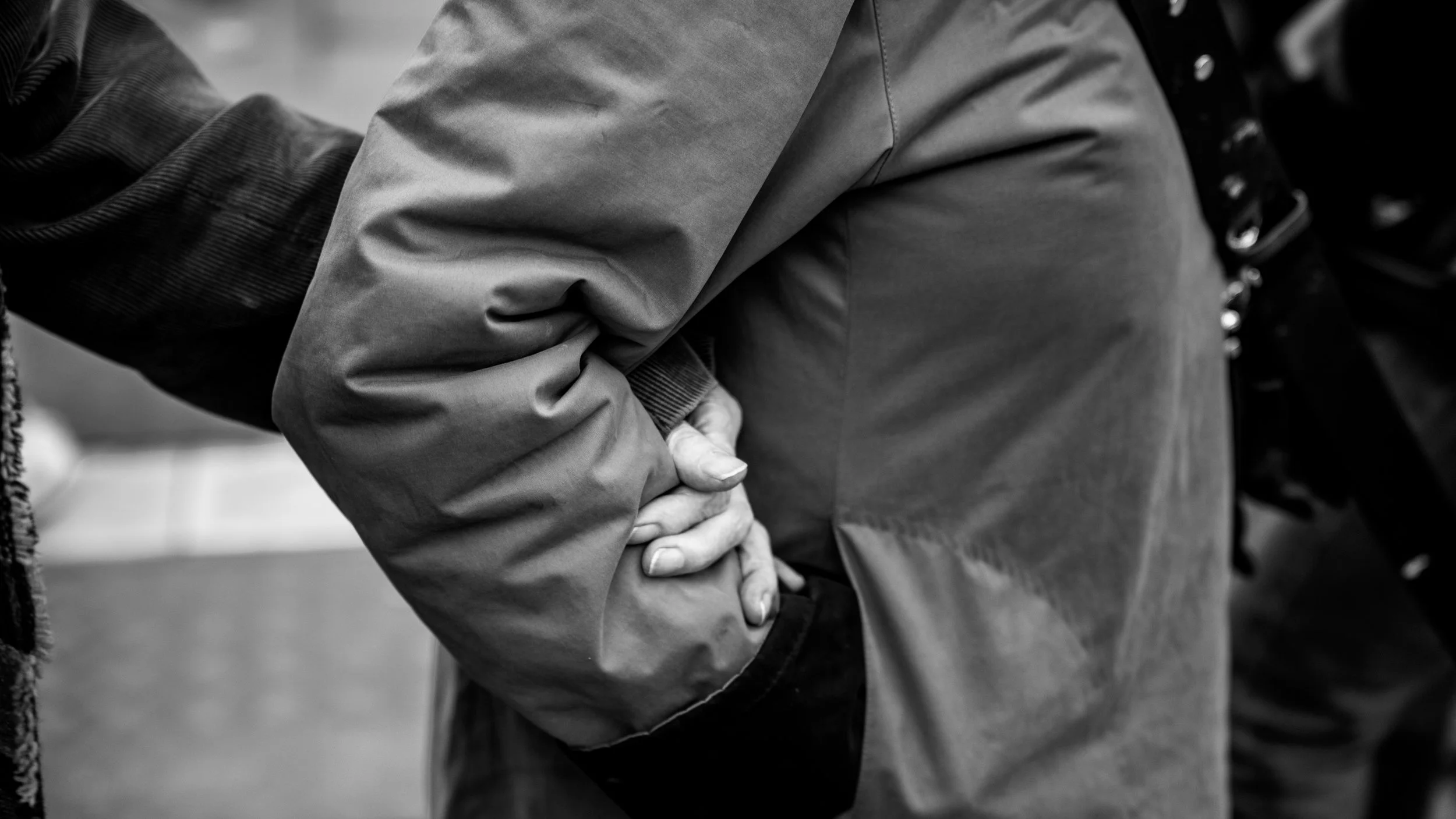 Close-up of a person wearing a jacket, with another person's hand grasping their wrist, suggesting a handshake or an arm-wrestling gesture.