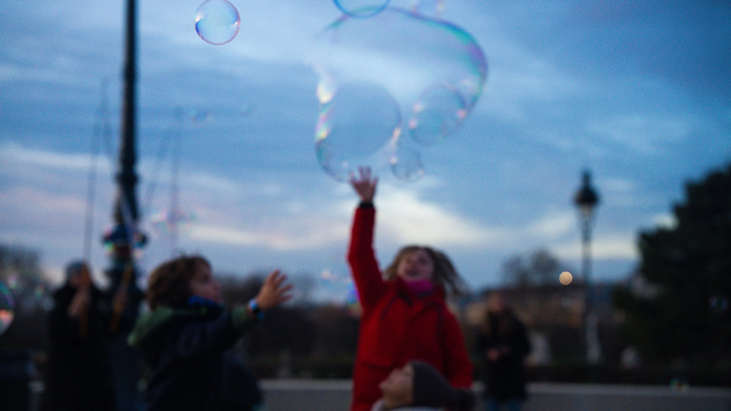 Children playing with soap bubbles outdoors during dusk, with a cloudy sky and streetlamp in the background.