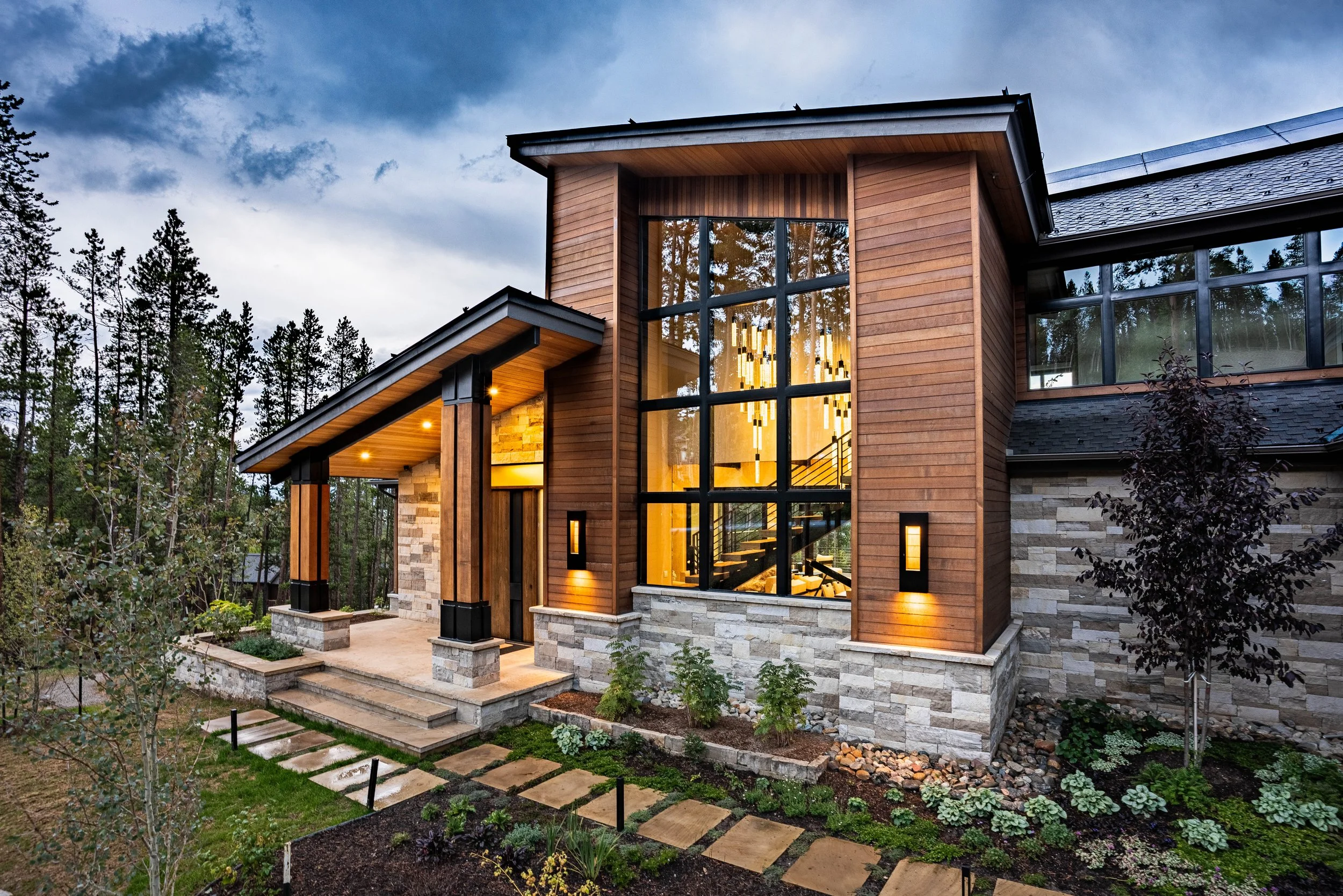 Modern house with large glass windows, wood siding, and stone accents, surrounded by a landscaped garden and trees.