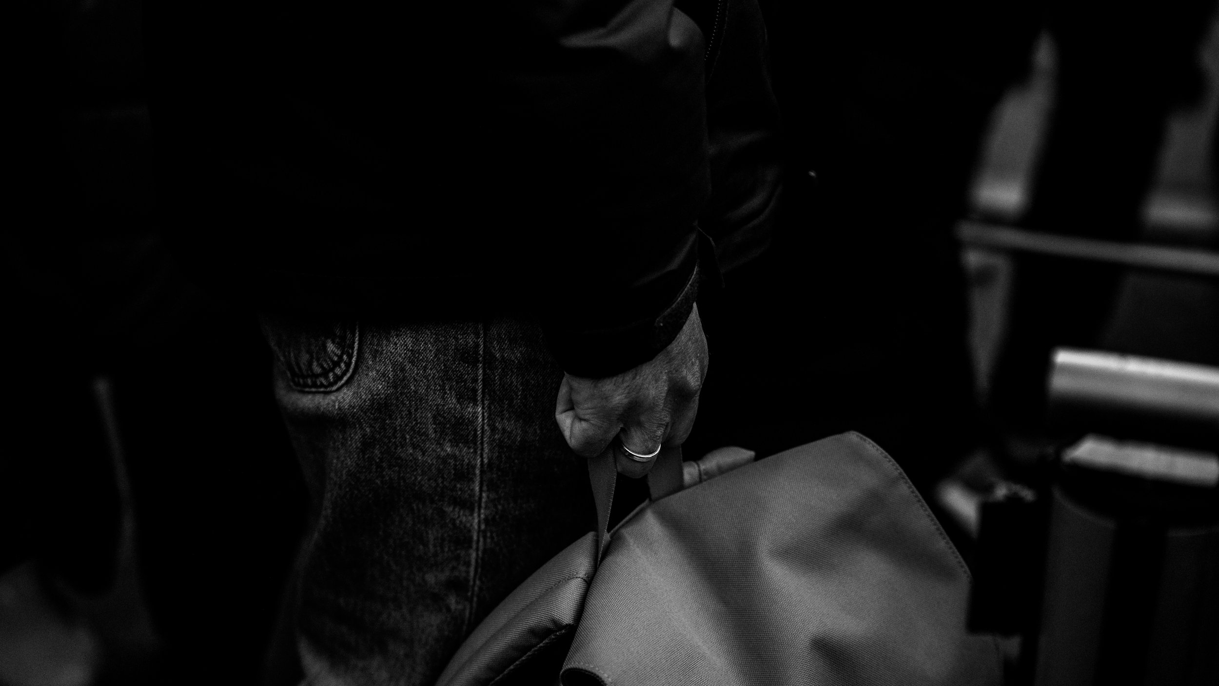 A person wearing jeans and a black jacket is holding a bag by its handle in a dark setting.