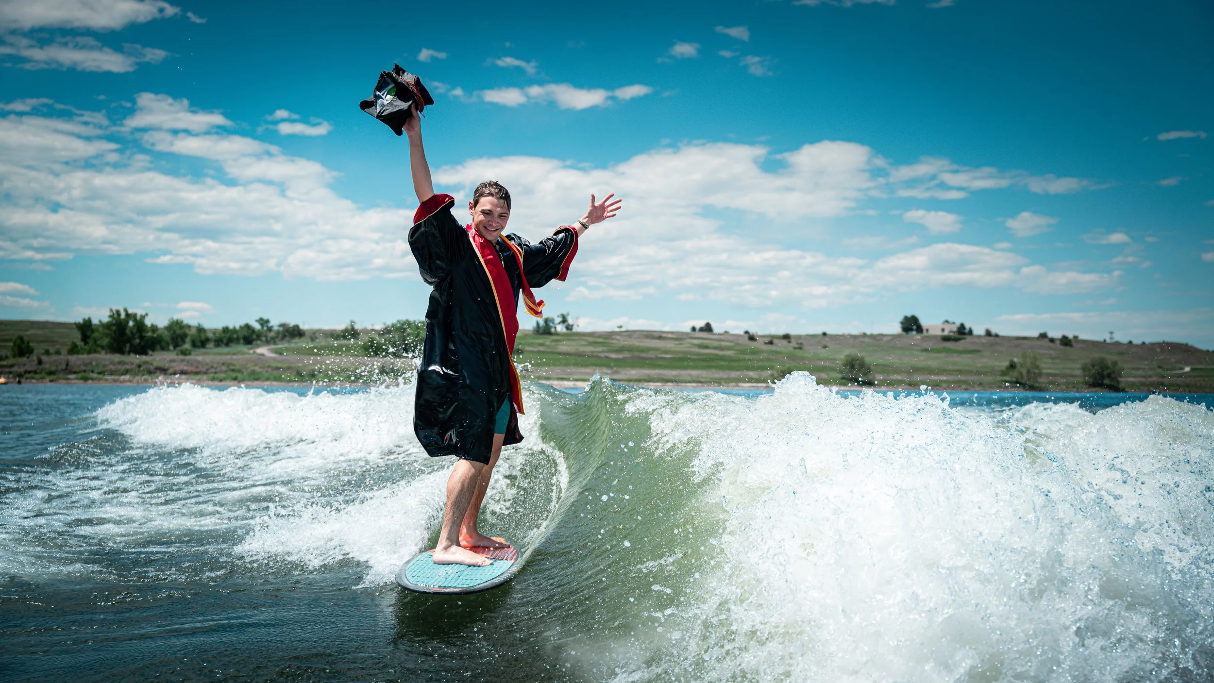 Person in a black and red graduation gown surfing on a wave in a large body of water under a partly cloudy sky, smiling and raising one arm.