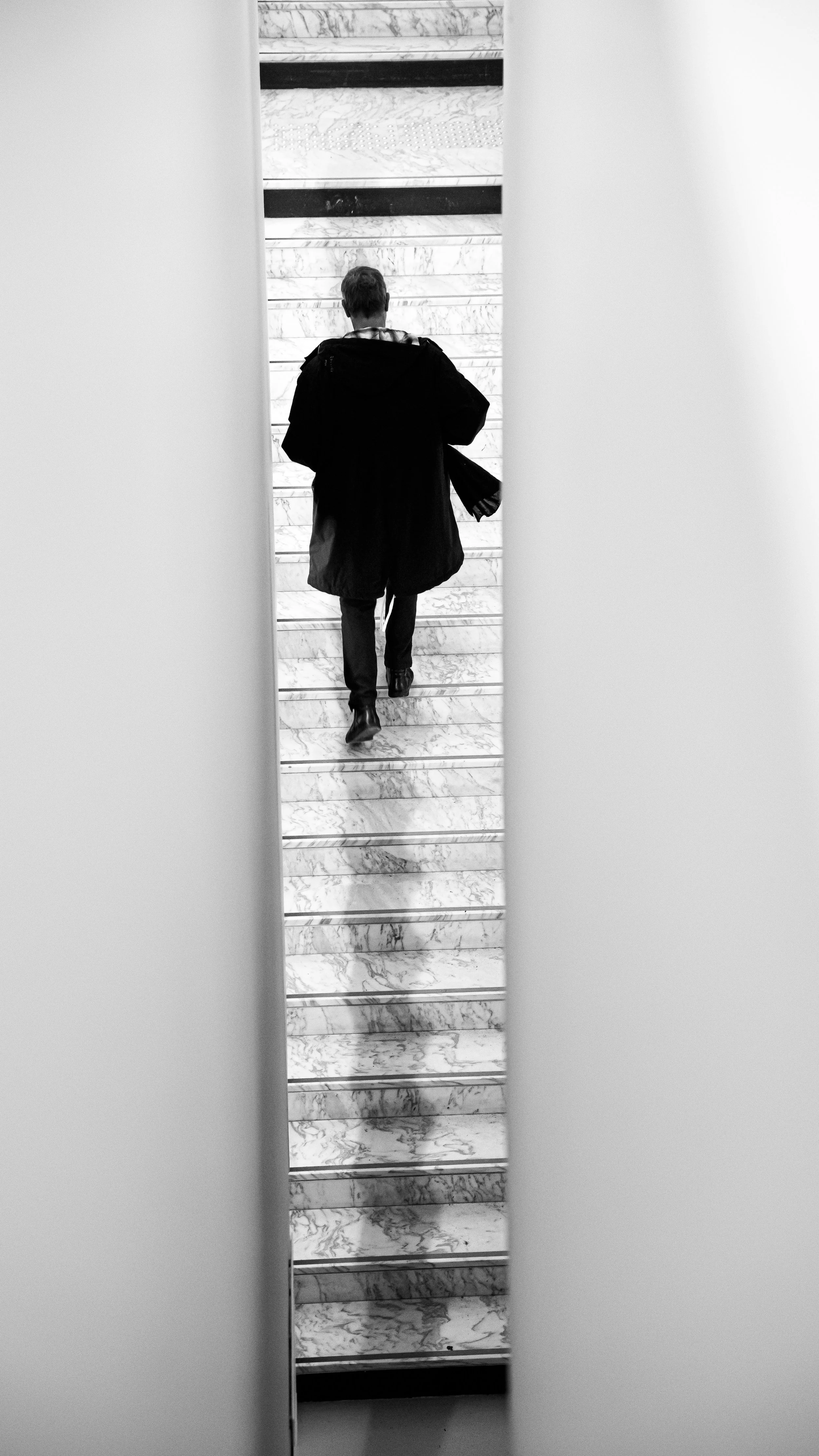 A person walking up marble stairs, seen through a narrow vertical mirror or opening, in black and white.