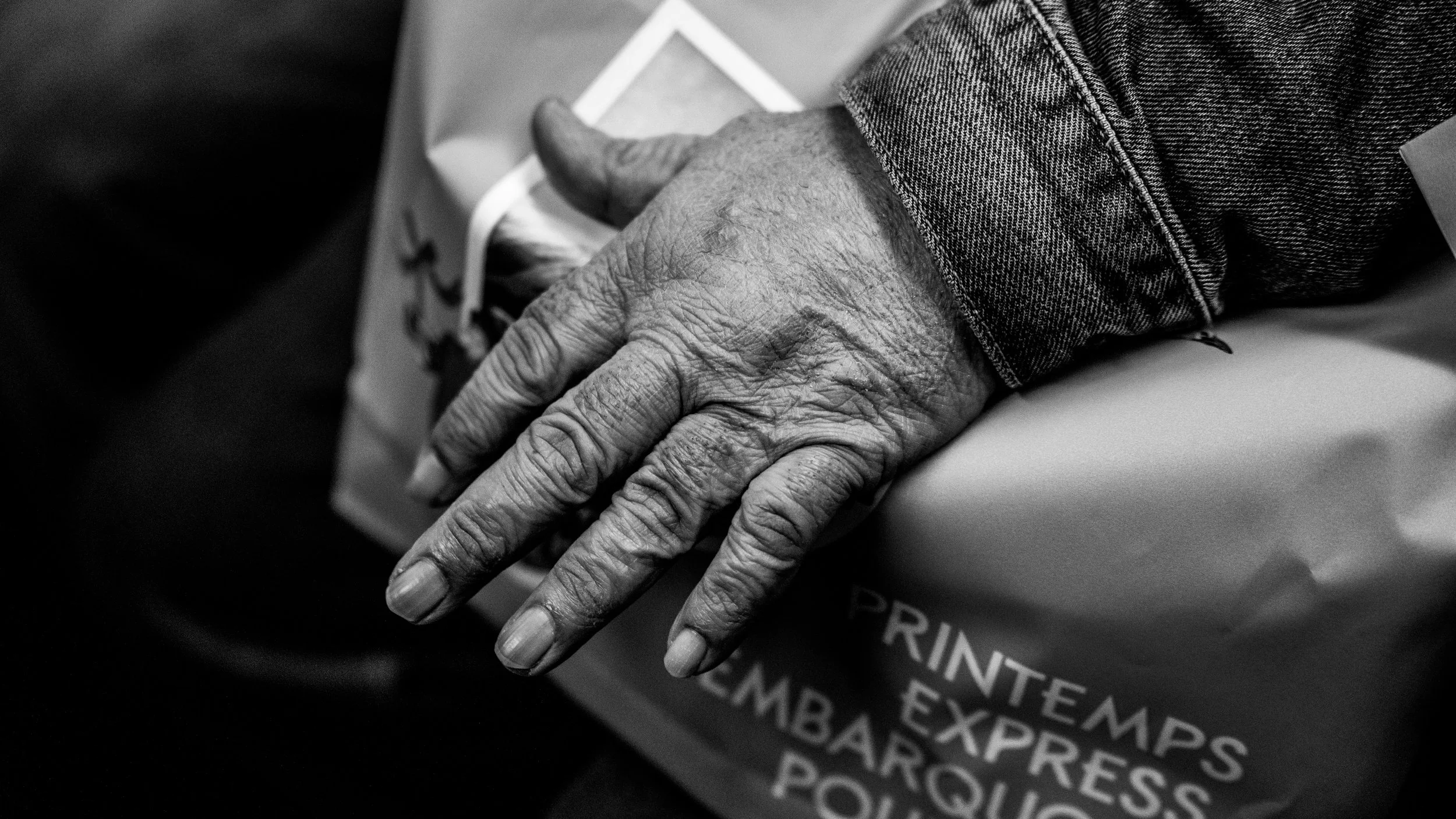 Close-up black and white photo of an elderly person's twisted, wrinkled hand resting on a paper bag with printed text, wearing a denim jacket.