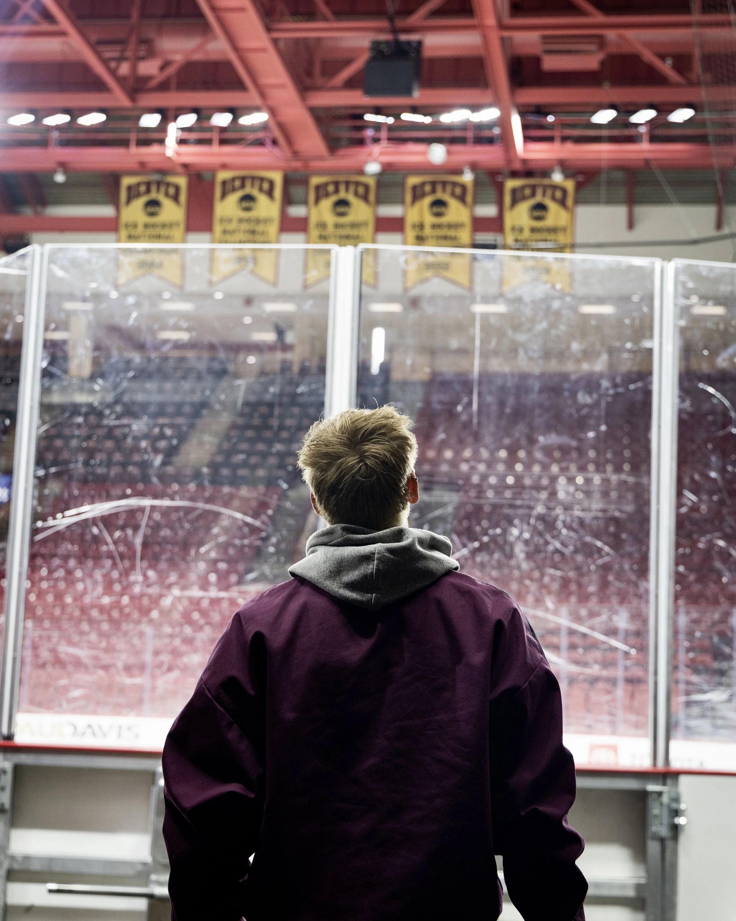 A person with blonde hair, wearing a maroon jacket and gray hoodie, looking at a damaged glass barrier on an ice rink inside a sports arena.