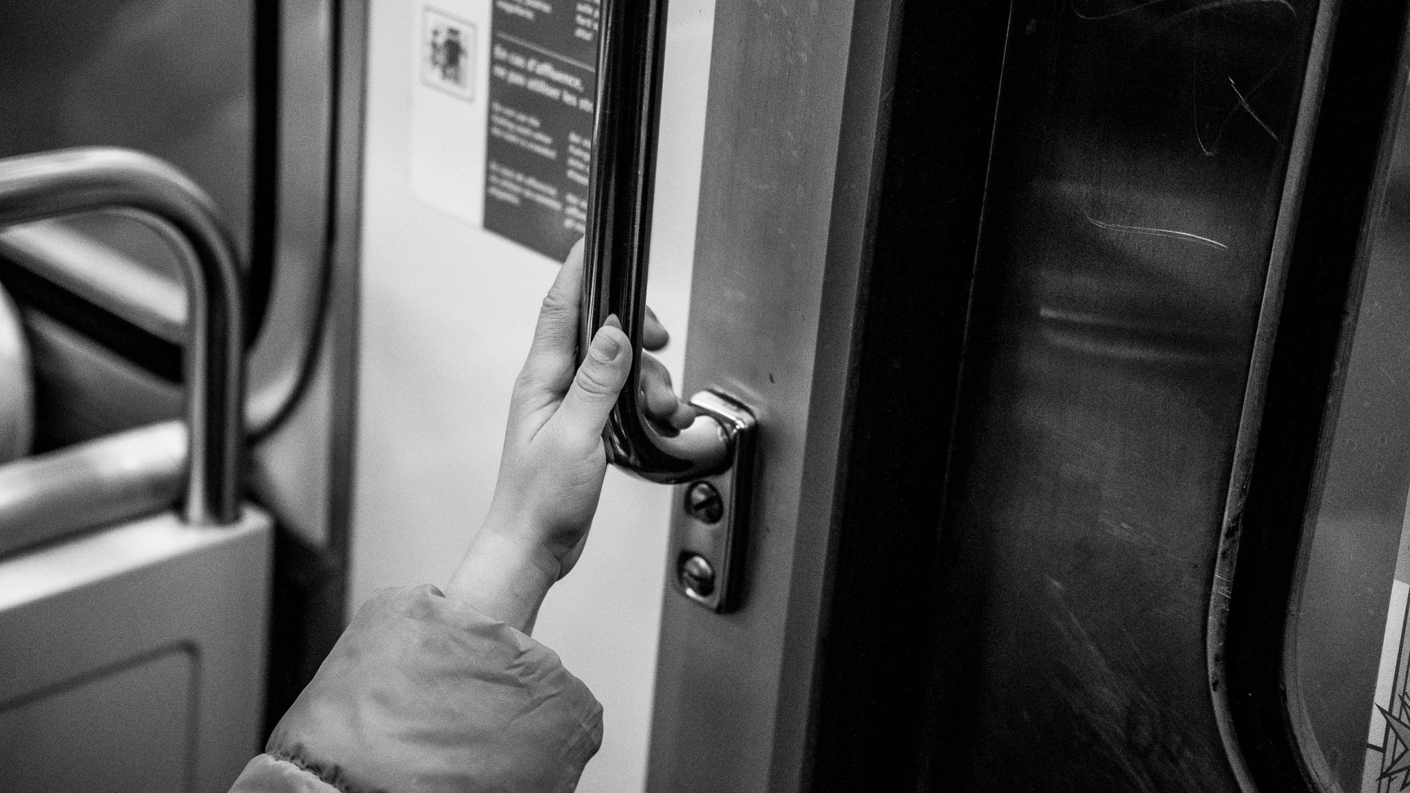 A person with a jacket is opening a door with a handle, likely on a bus or train, in black and white.
