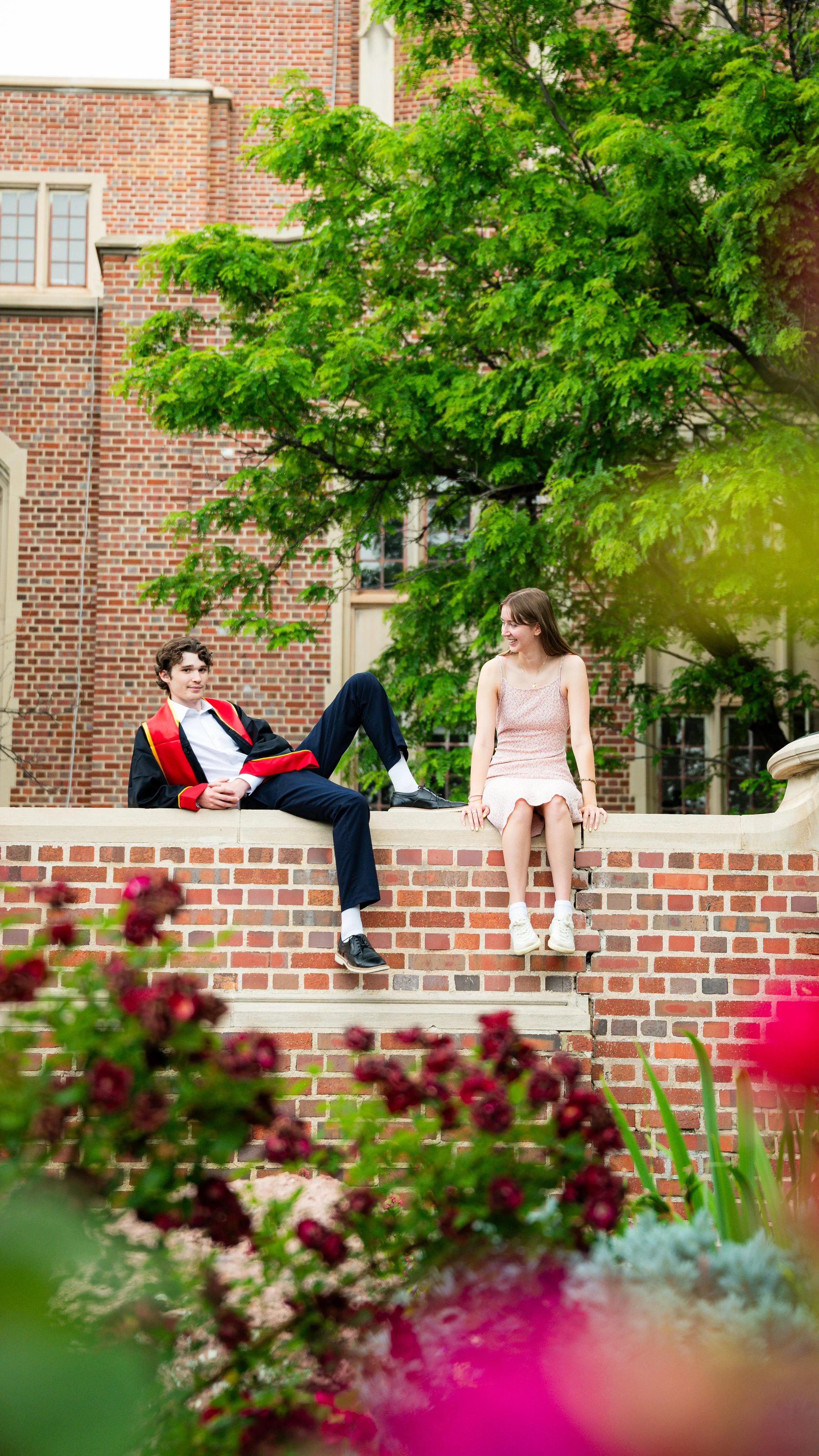 A young man in a graduation gown and a young woman in a dress sit on a brick wall outdoors, smiling and looking at each other, with greenery and a brick building in the background.