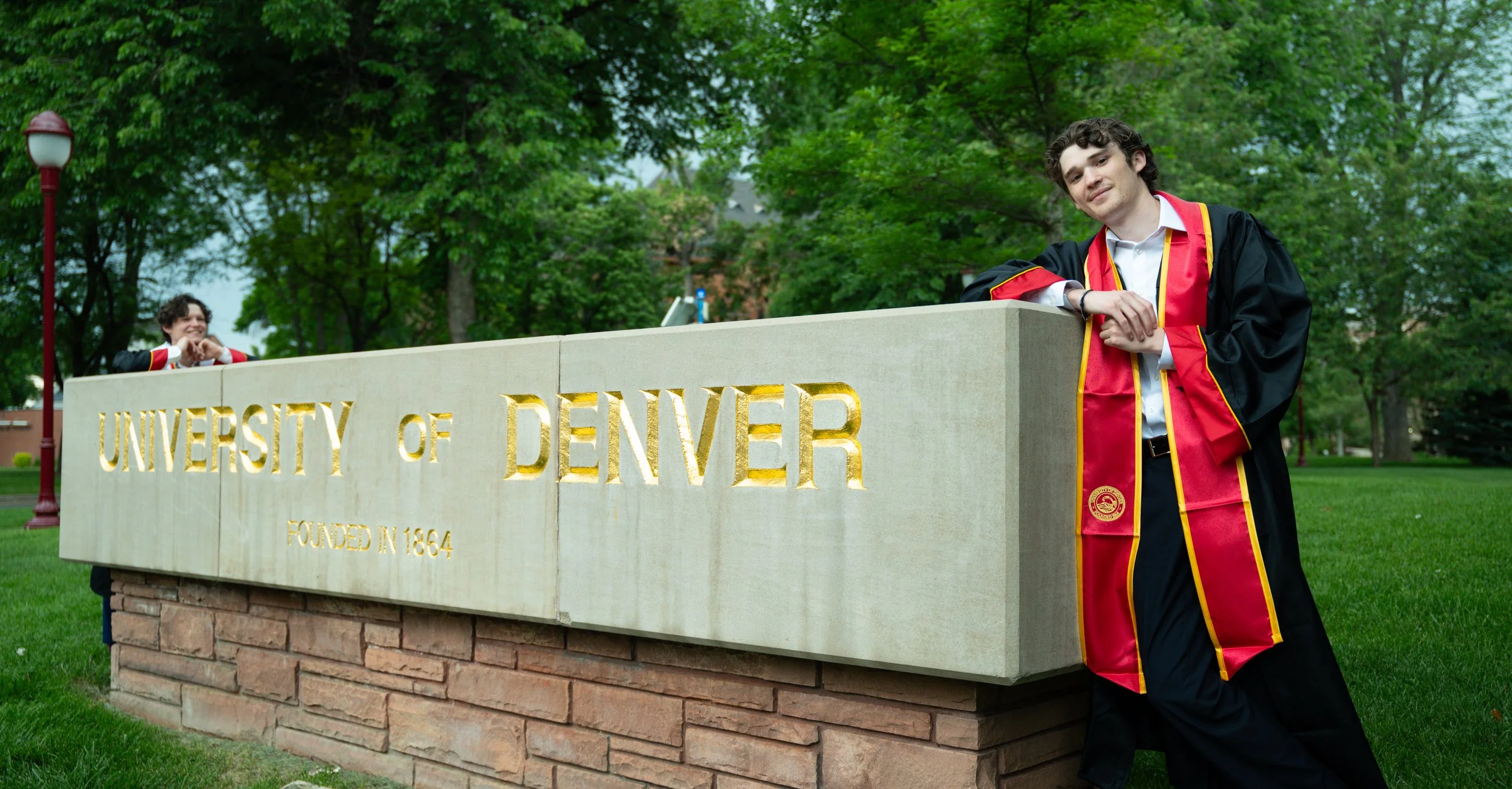 A young man in graduation attire leaning on a stone sign that reads "University of Denver Founded in 1864" with green trees in the background.