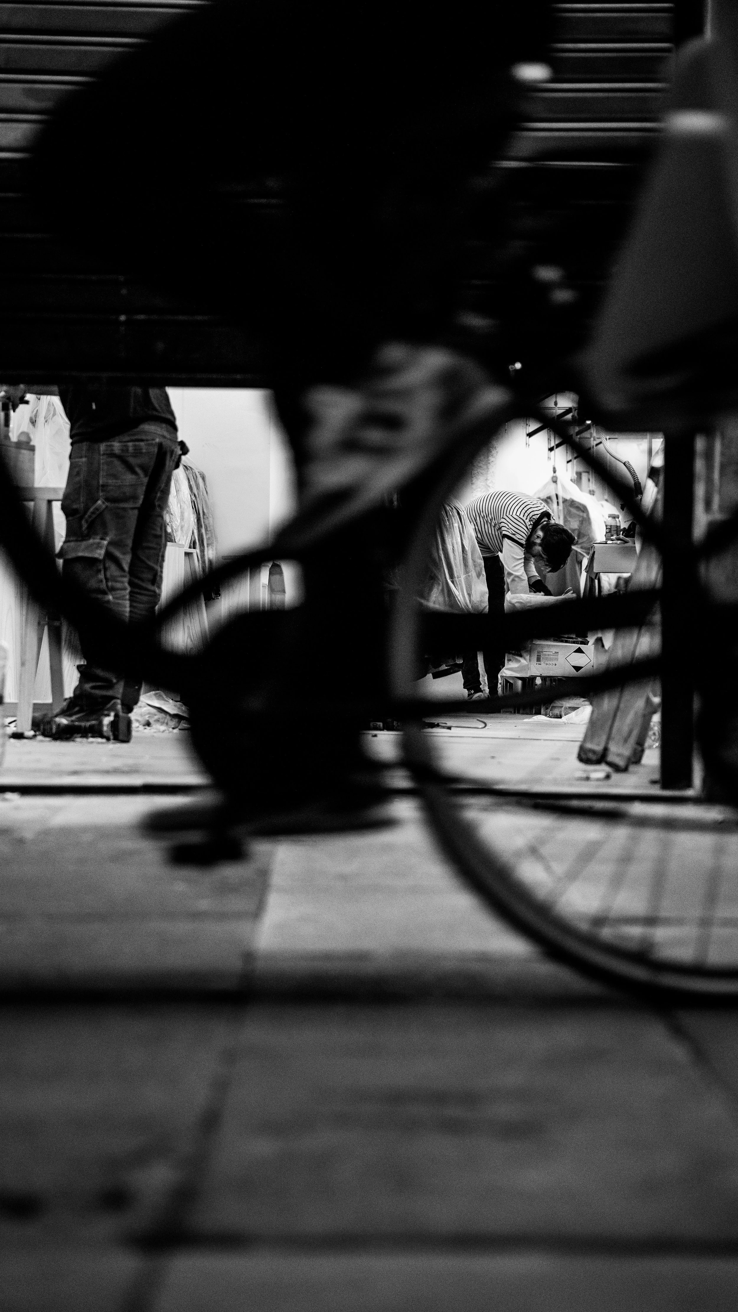 Black and white photo taken from a low angle showing people working at a workshop or studio, with a focus on a person bending over a table in the background.
