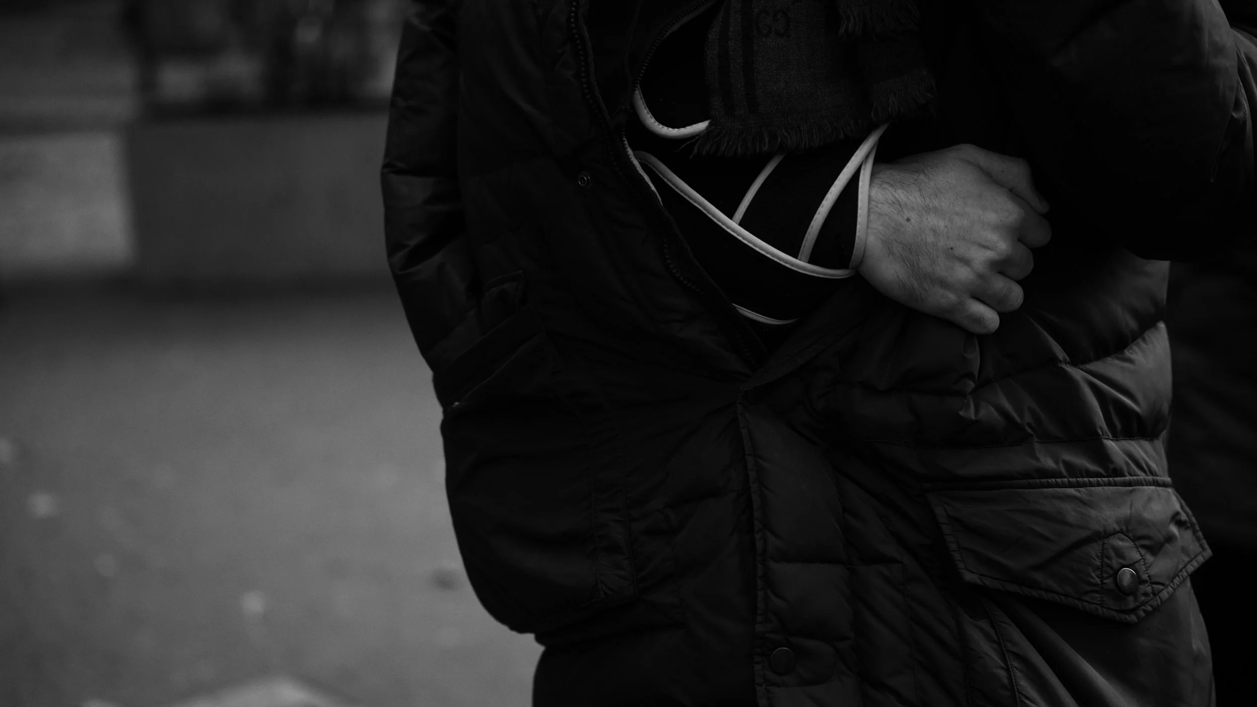 Black and white photo of a person wearing a dark puffer jacket and a scarf, clutching their chest with one hand, with blurred outdoor background.