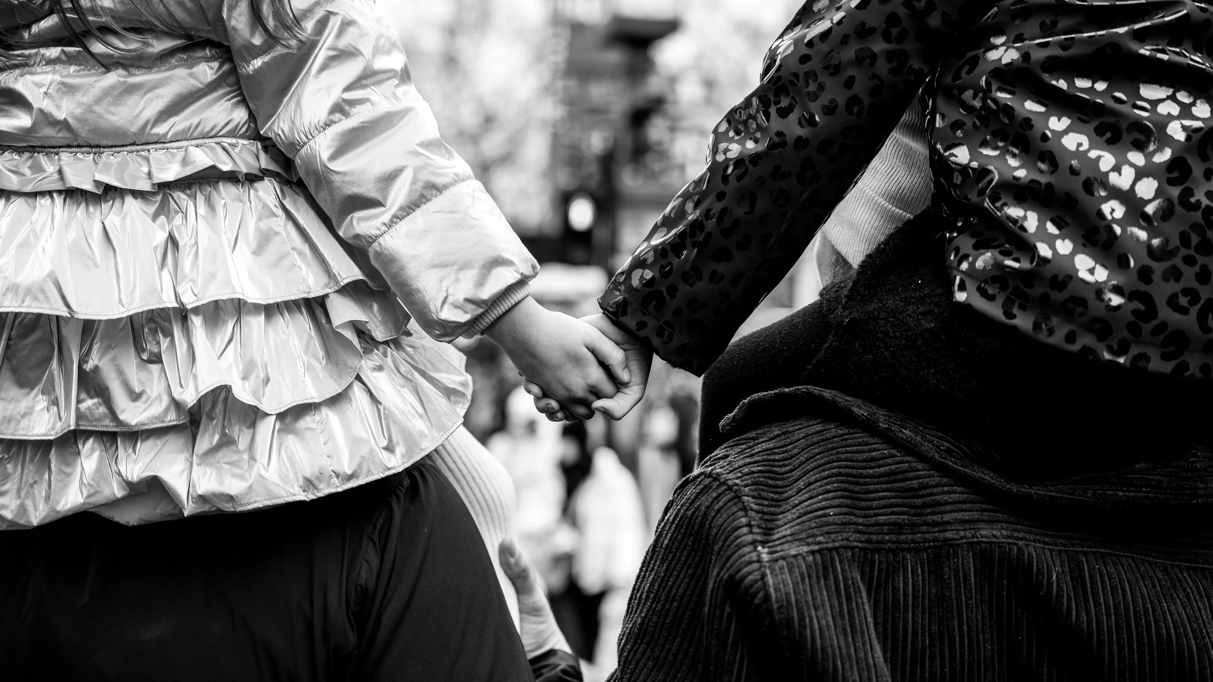 Two children holding hands, one wearing a shiny jacket and the other a patterned jacket, in a black and white photo.