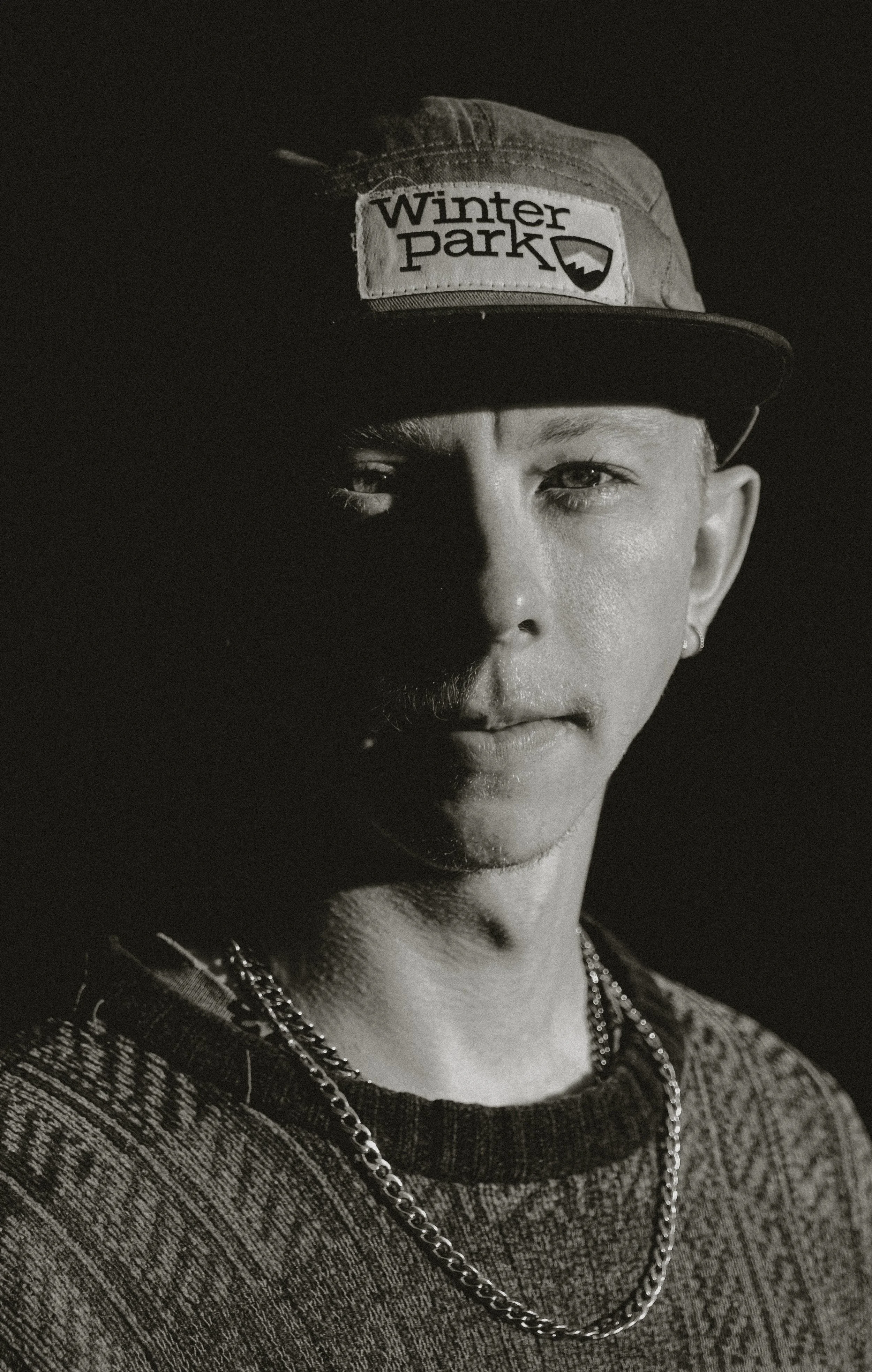Black and white portrait of a young person wearing a baseball cap with 'Winter Park' logo, looking directly at the camera with one side of face shadowed, wearing a patterned sweater and chain necklace.