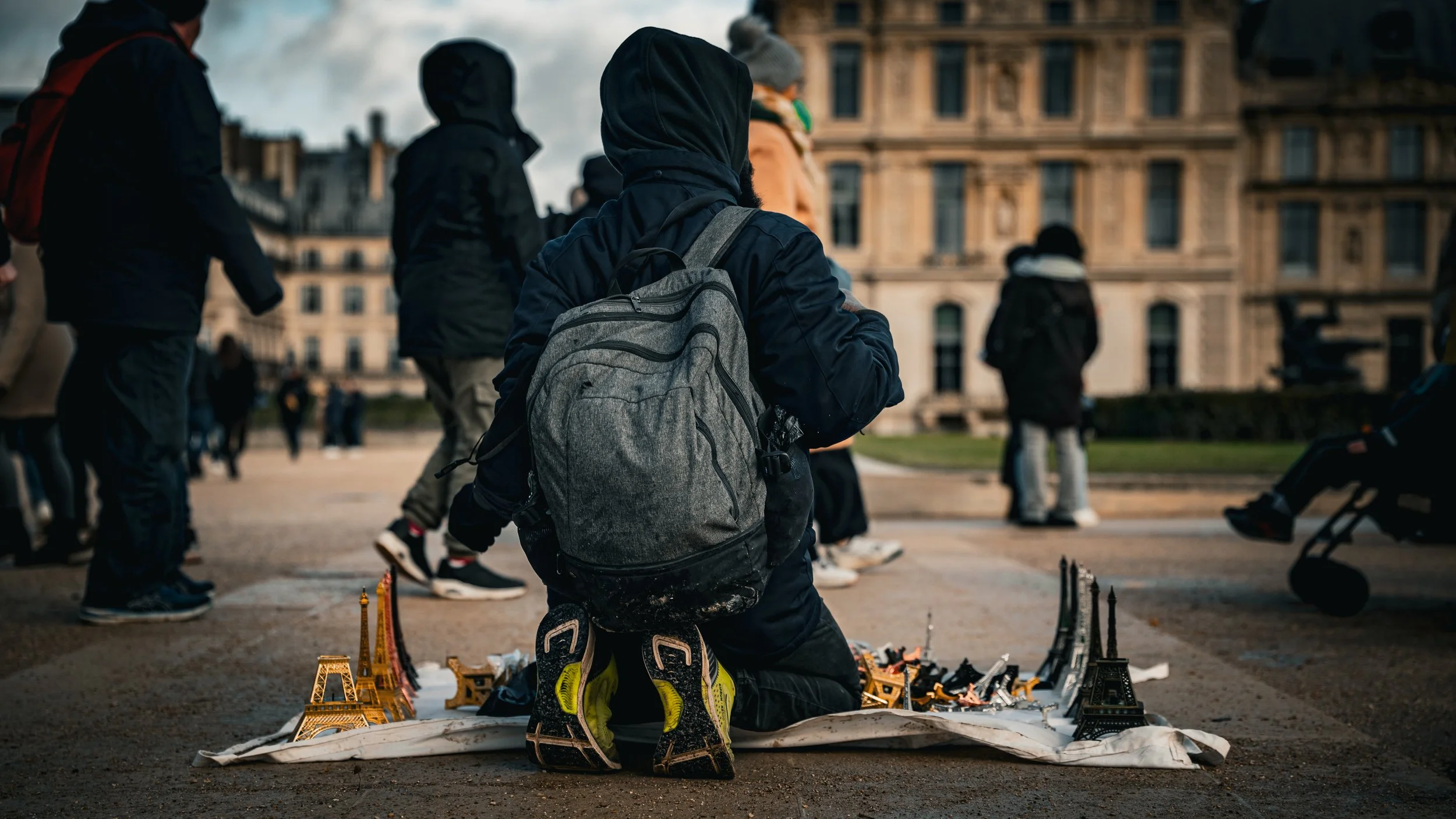 A person kneels on the ground at a street market in front of small Eiffel Tower replicas for sale, with other pedestrians walking by in a city.