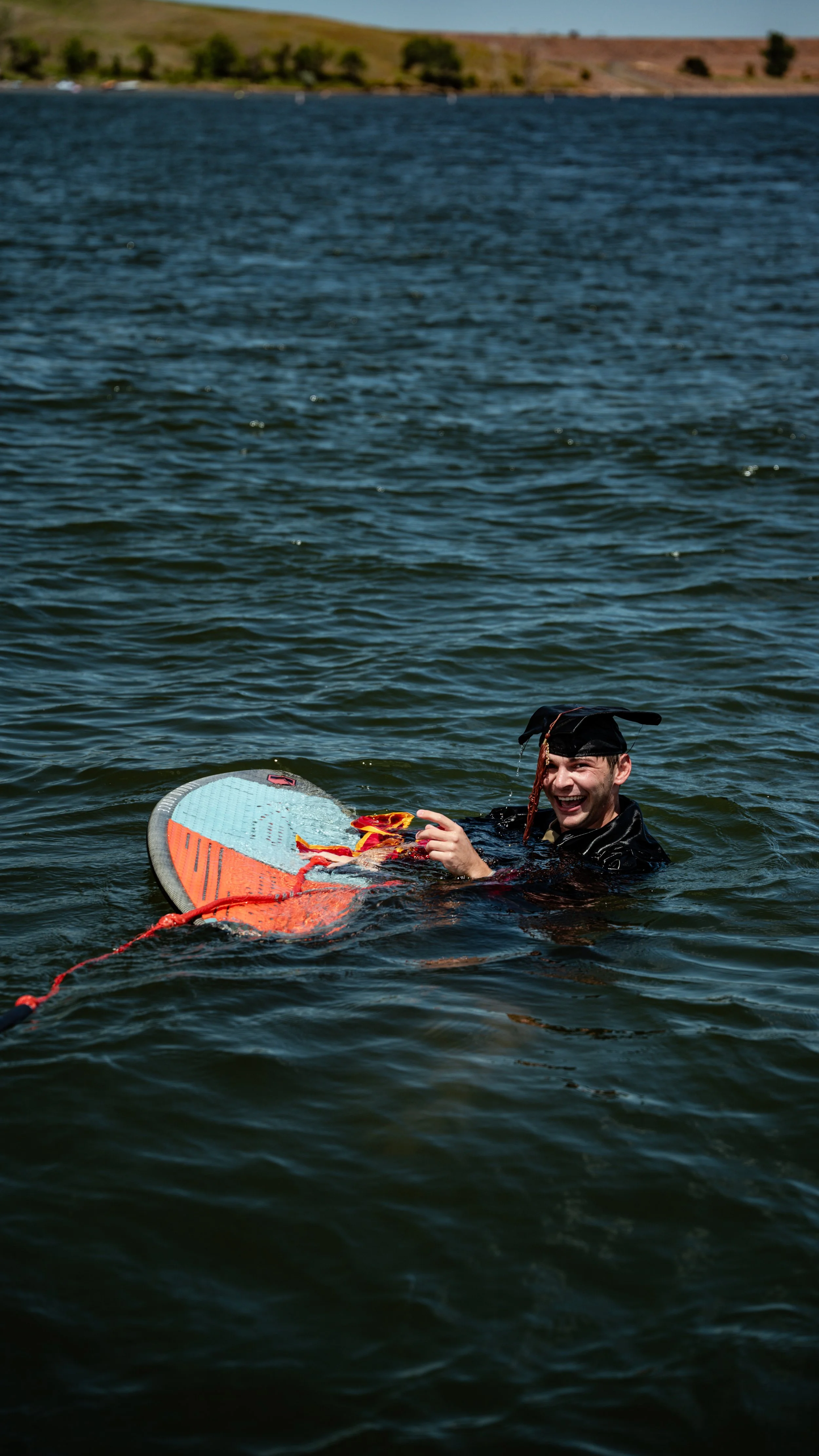 A man in a graduation gown and cap swimming in a lake, holding a colorful surfboard, smiling and enjoying the water.