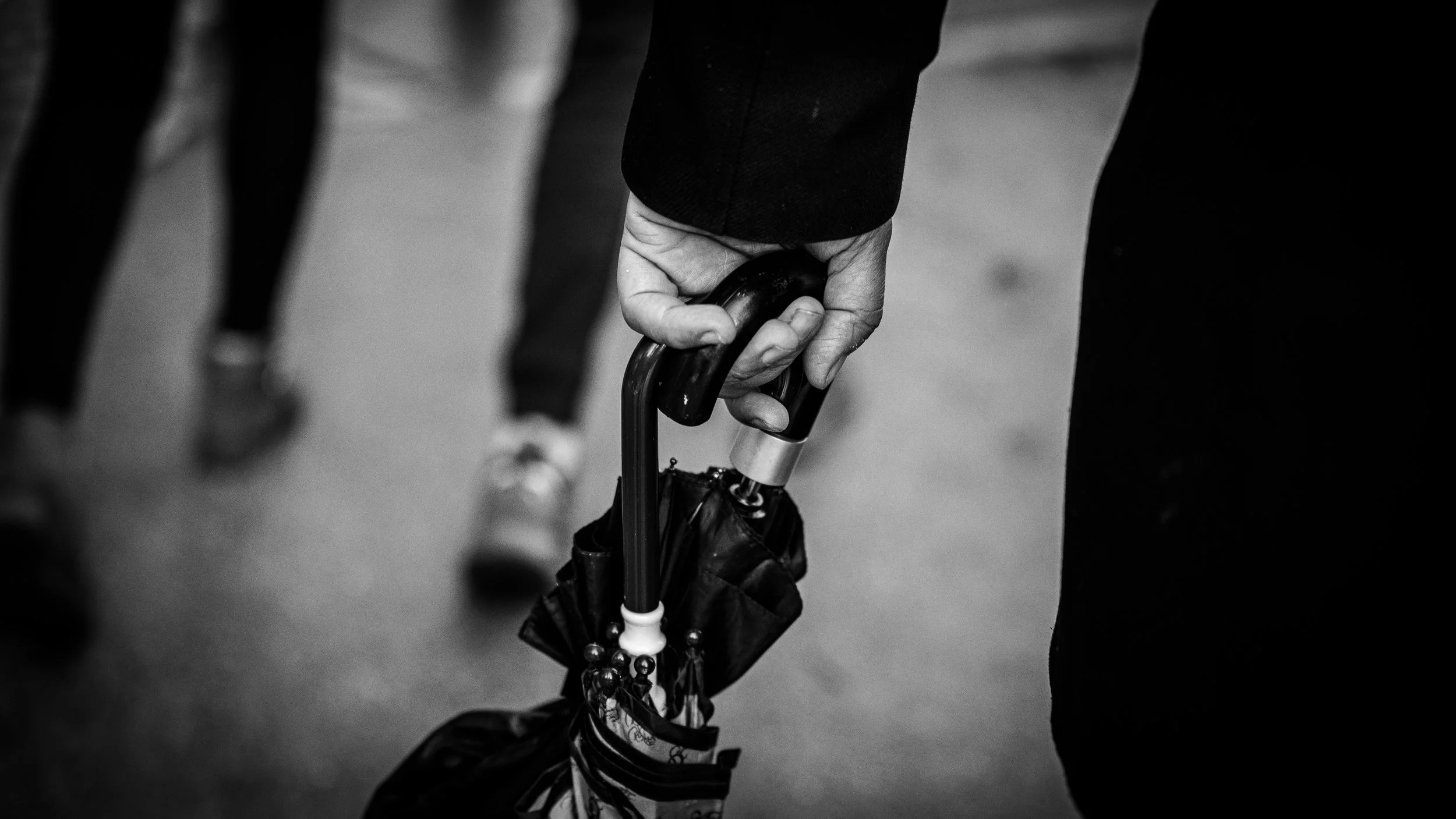 Close-up of a person's hand holding a black umbrella with a curved handle, with other people's legs visible in the background.