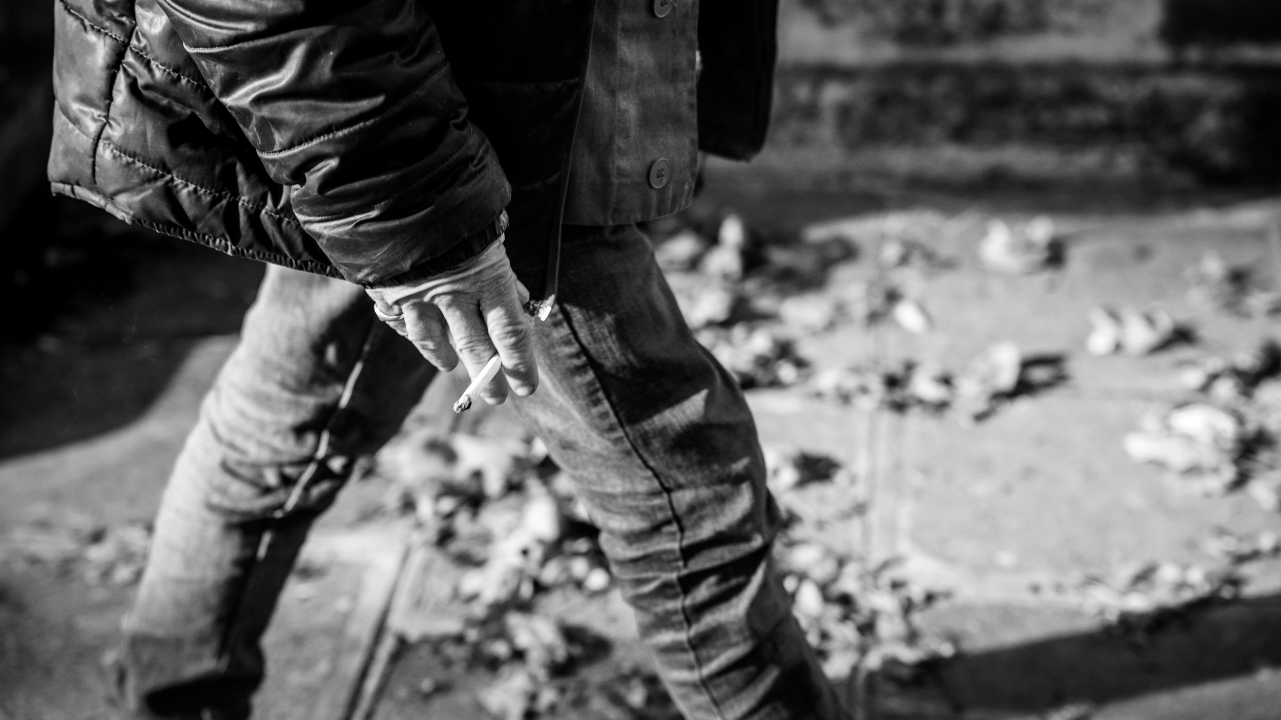 Person wearing a jacket and jeans, holding a cigarette in their right hand, walking outdoors on a sidewalk with fallen leaves.