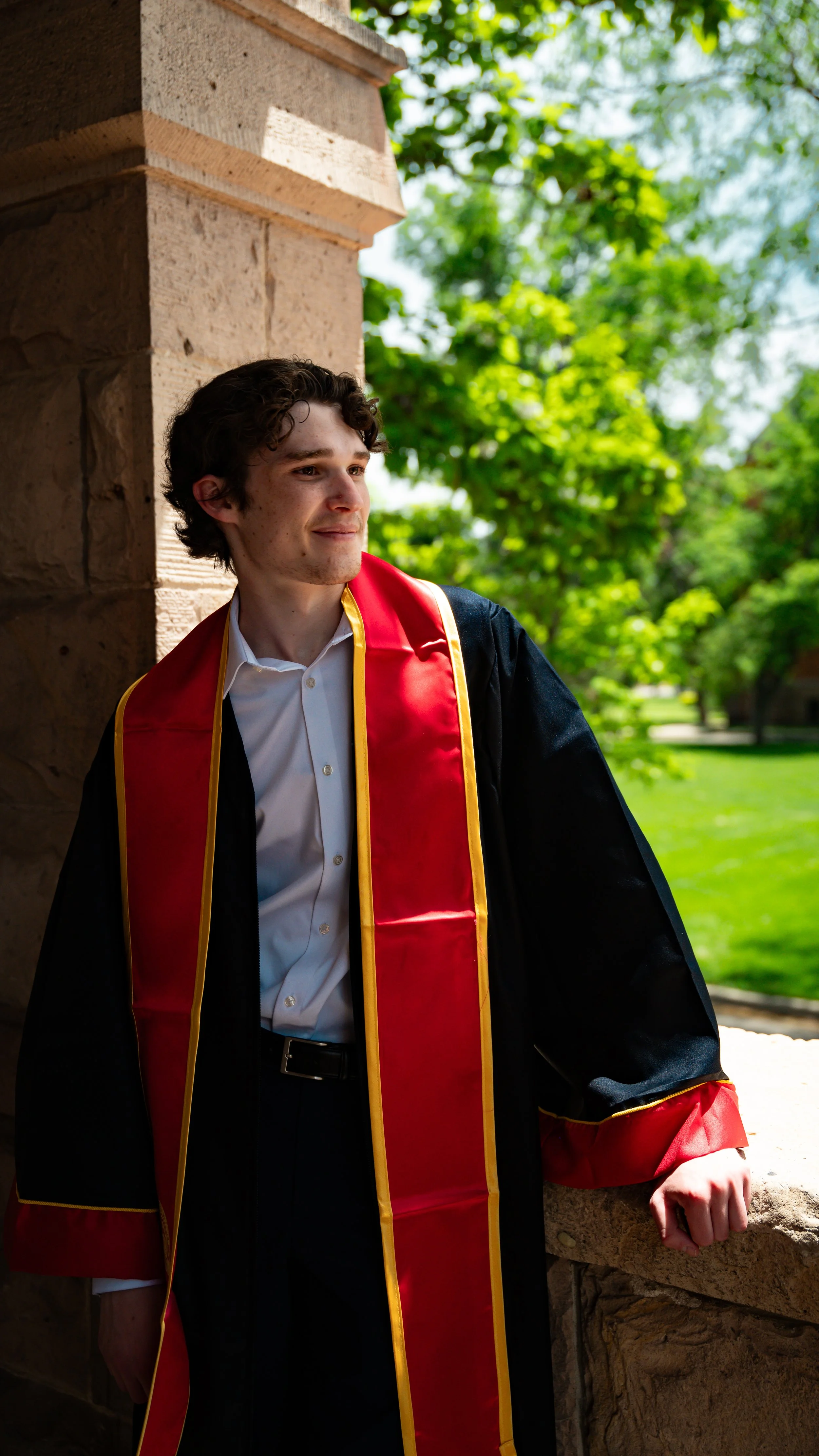 Young man in graduation gown with red and gold sash, standing outside by a stone railing, with trees and greenery in the background.