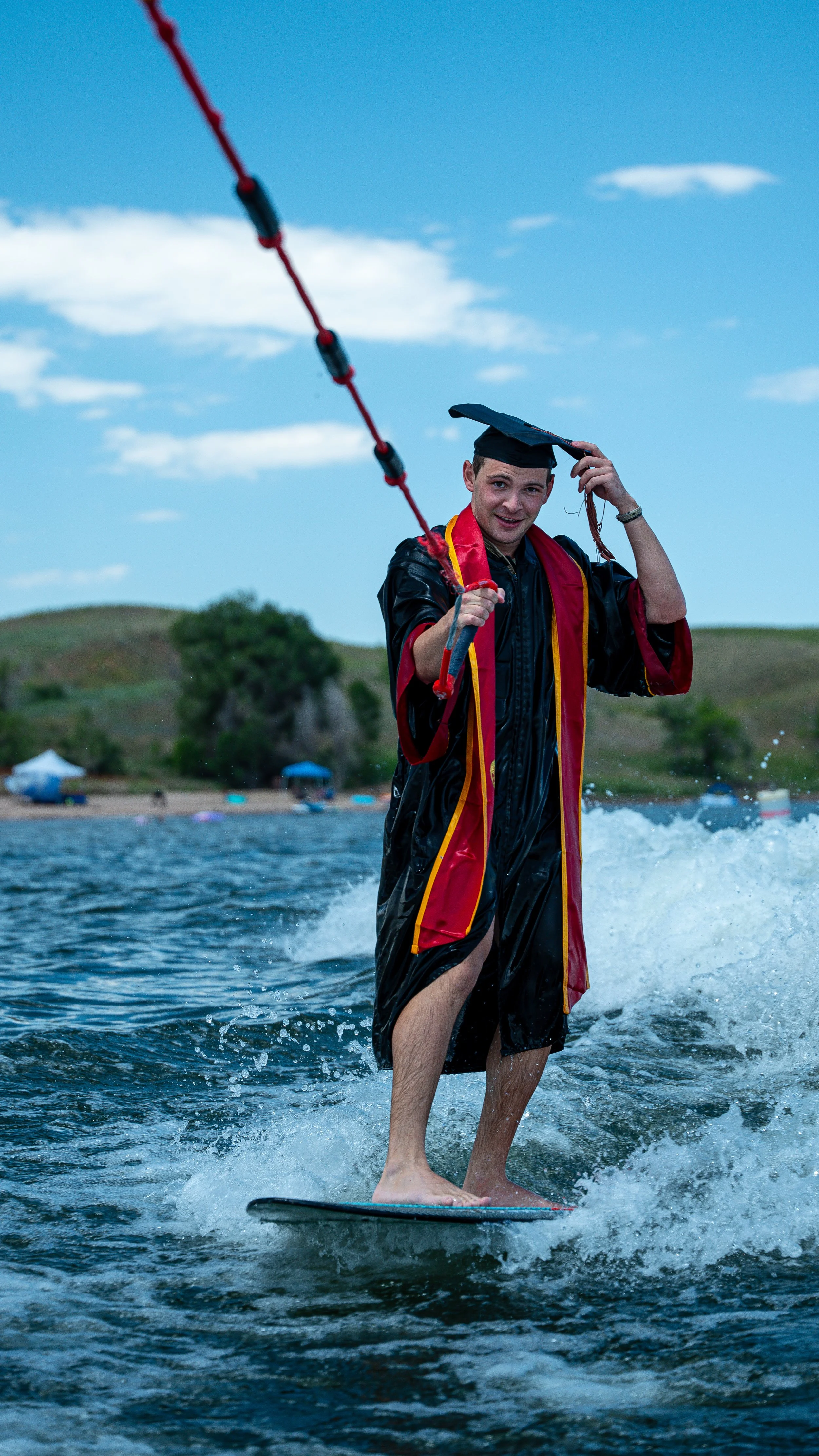 Young man in graduation gown and cap surfing on a lake with a shoreline and green hills in the background.