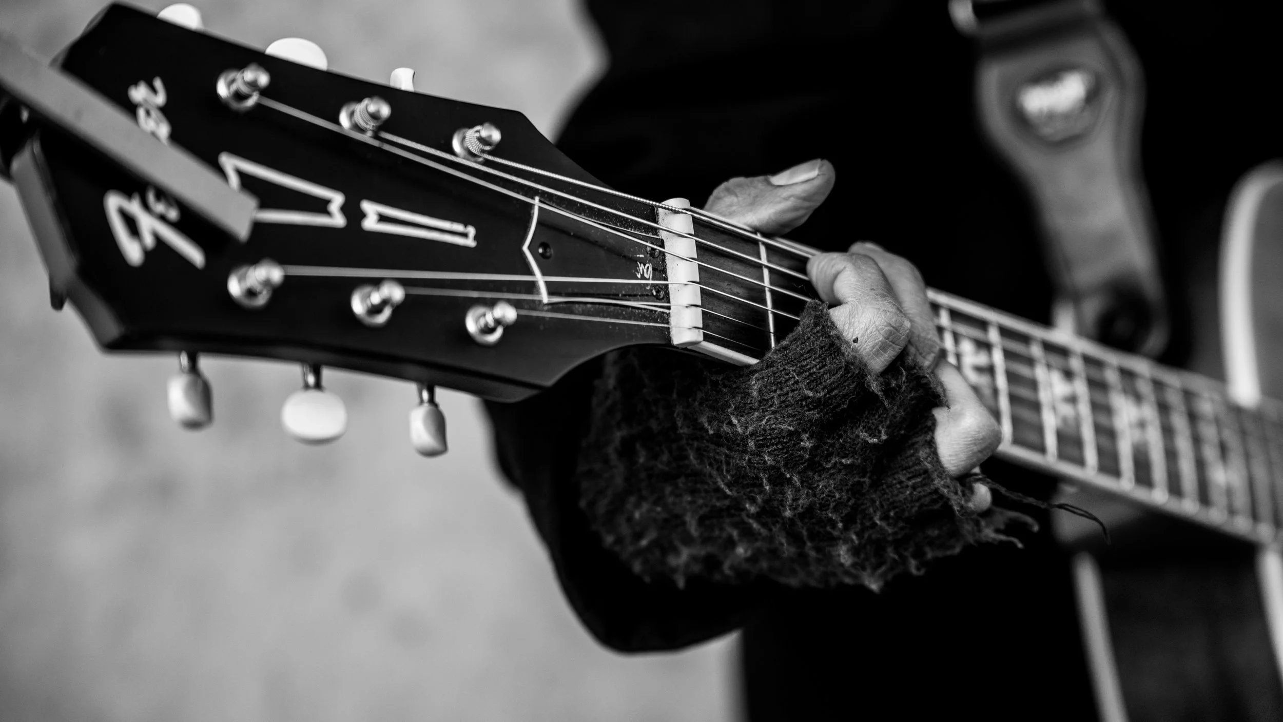 Close-up of a person playing an acoustic guitar, with focus on hand on fretboard and guitar headstock in black and white.