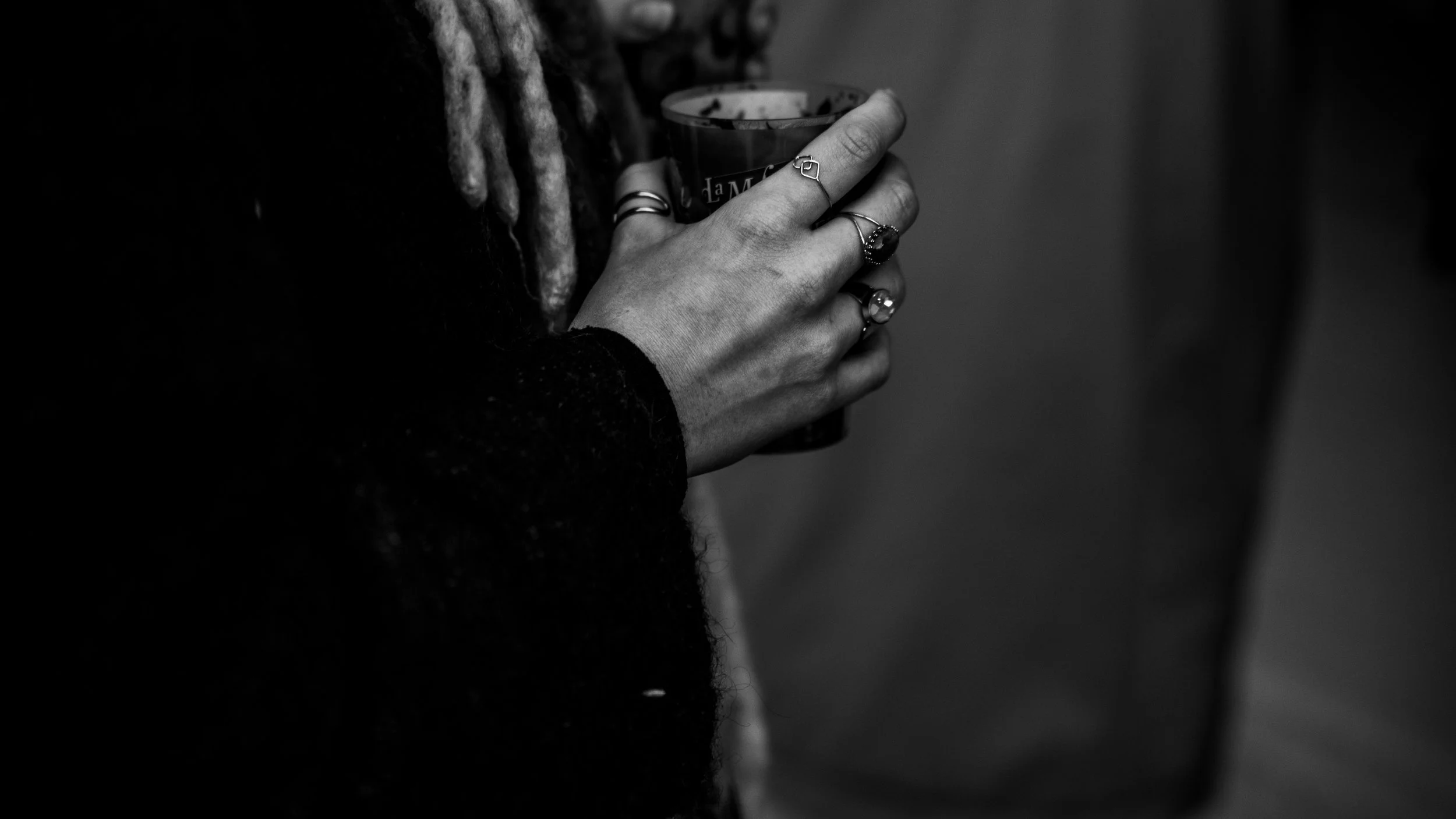 Close-up of a person's hand holding a cup, with multiple rings on fingers, in black and white.