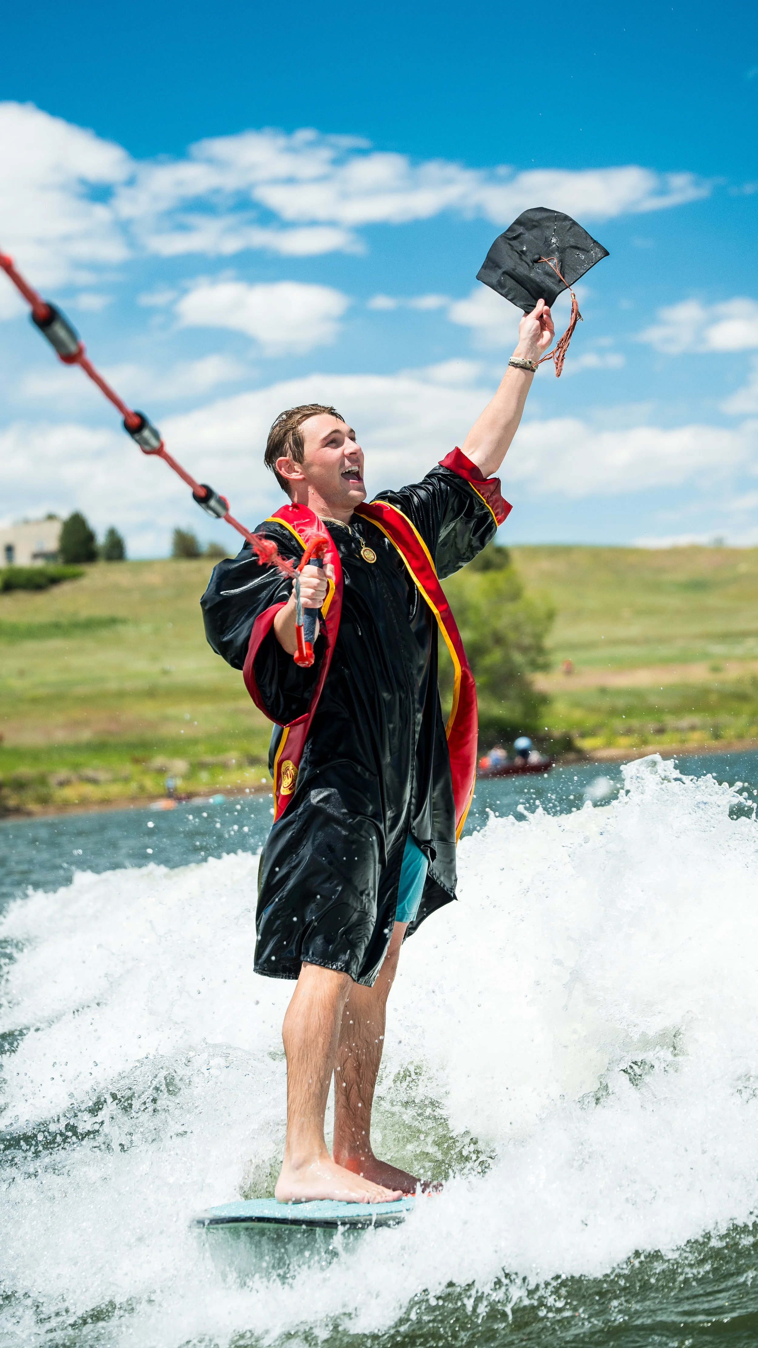 Young man wakes on a wakeboard, holding a graduation cap and a water ski rope, celebrating graduation on a lake during the day with a blue sky and clouds.