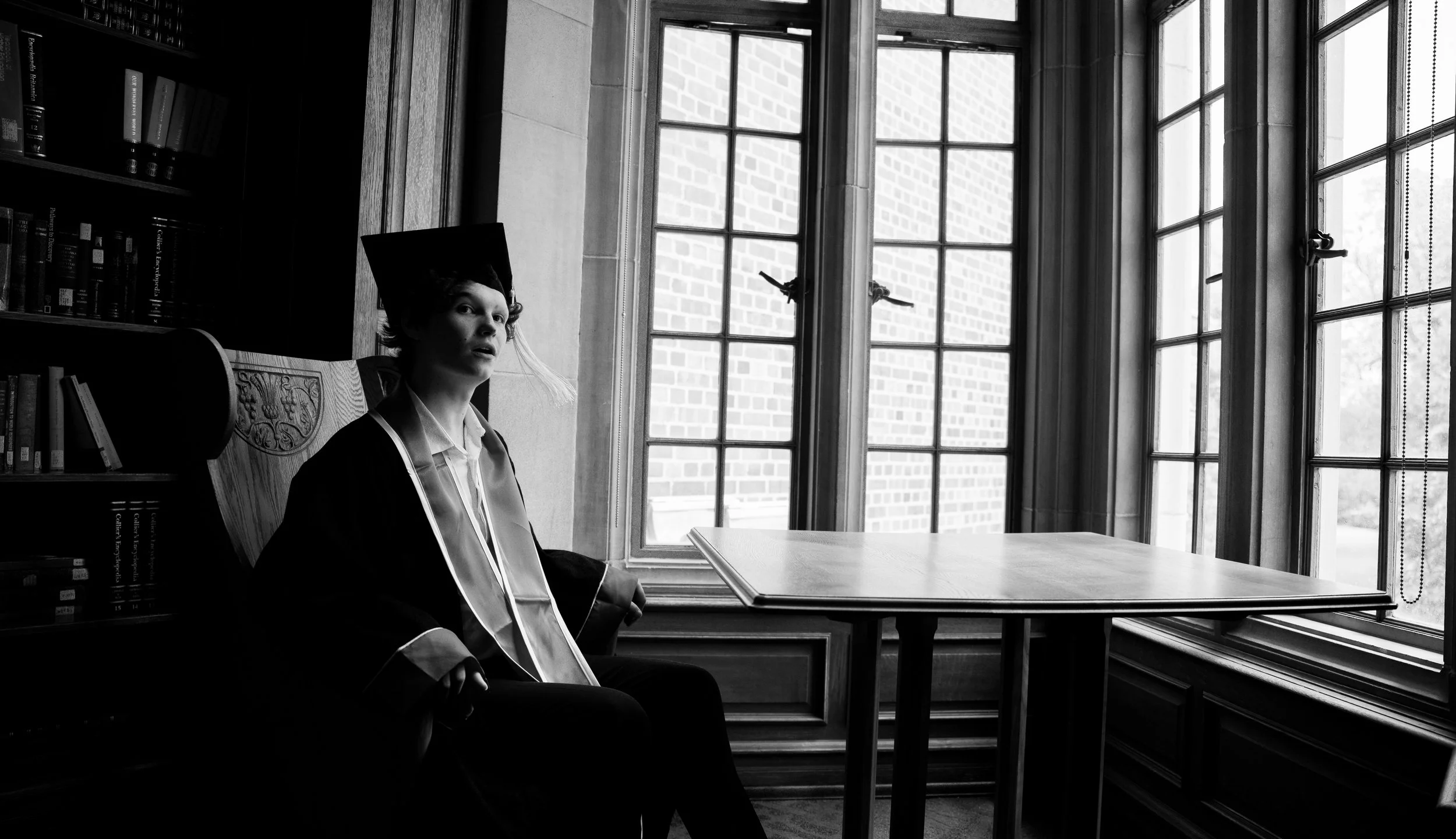 A person in graduation cap and gown sitting in a library room with large windows and brick exterior visible outside.