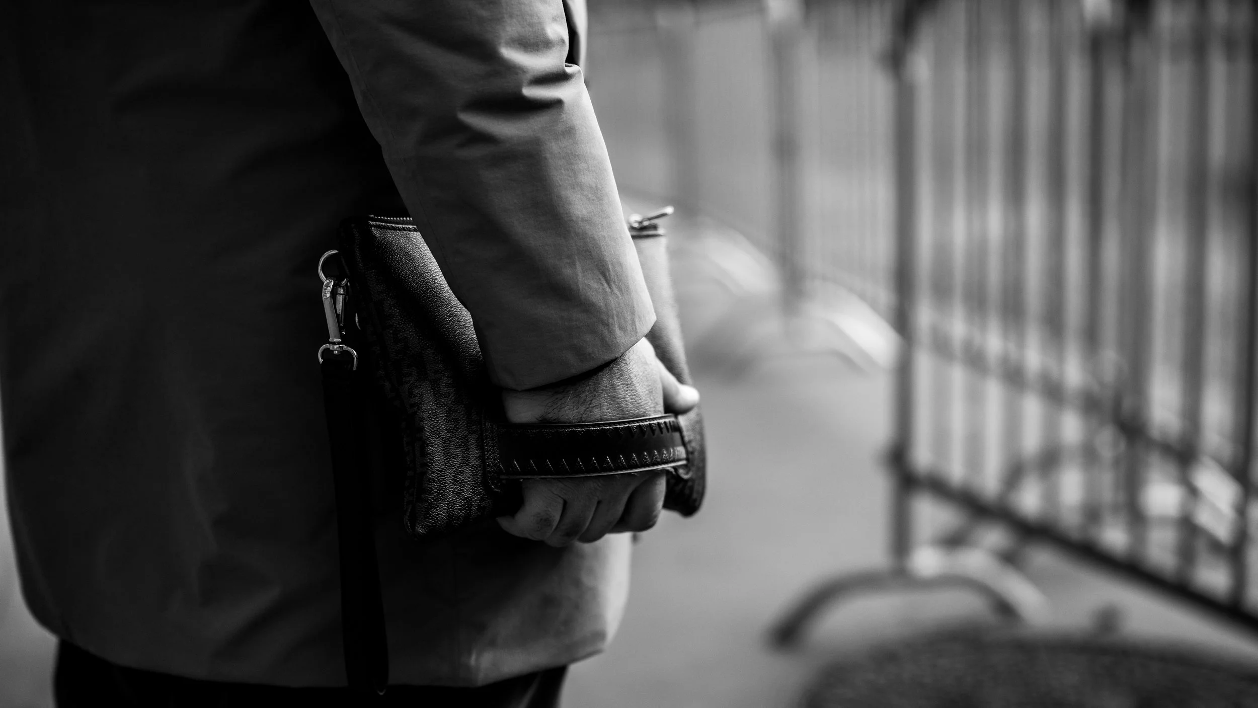 A person holding a wallet or clutch in their hand. The image is in black and white and appears to be in a public space with metal railings in the background.