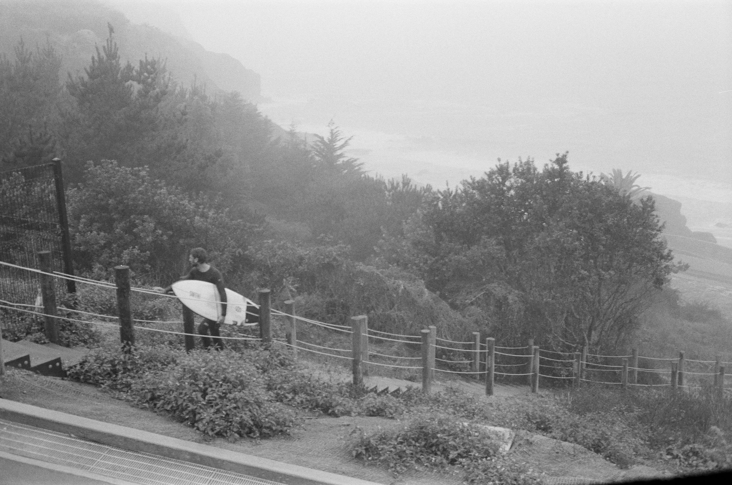 A person with a surfboard walking along a fenced pathway in a foggy, wooded area near the coast.