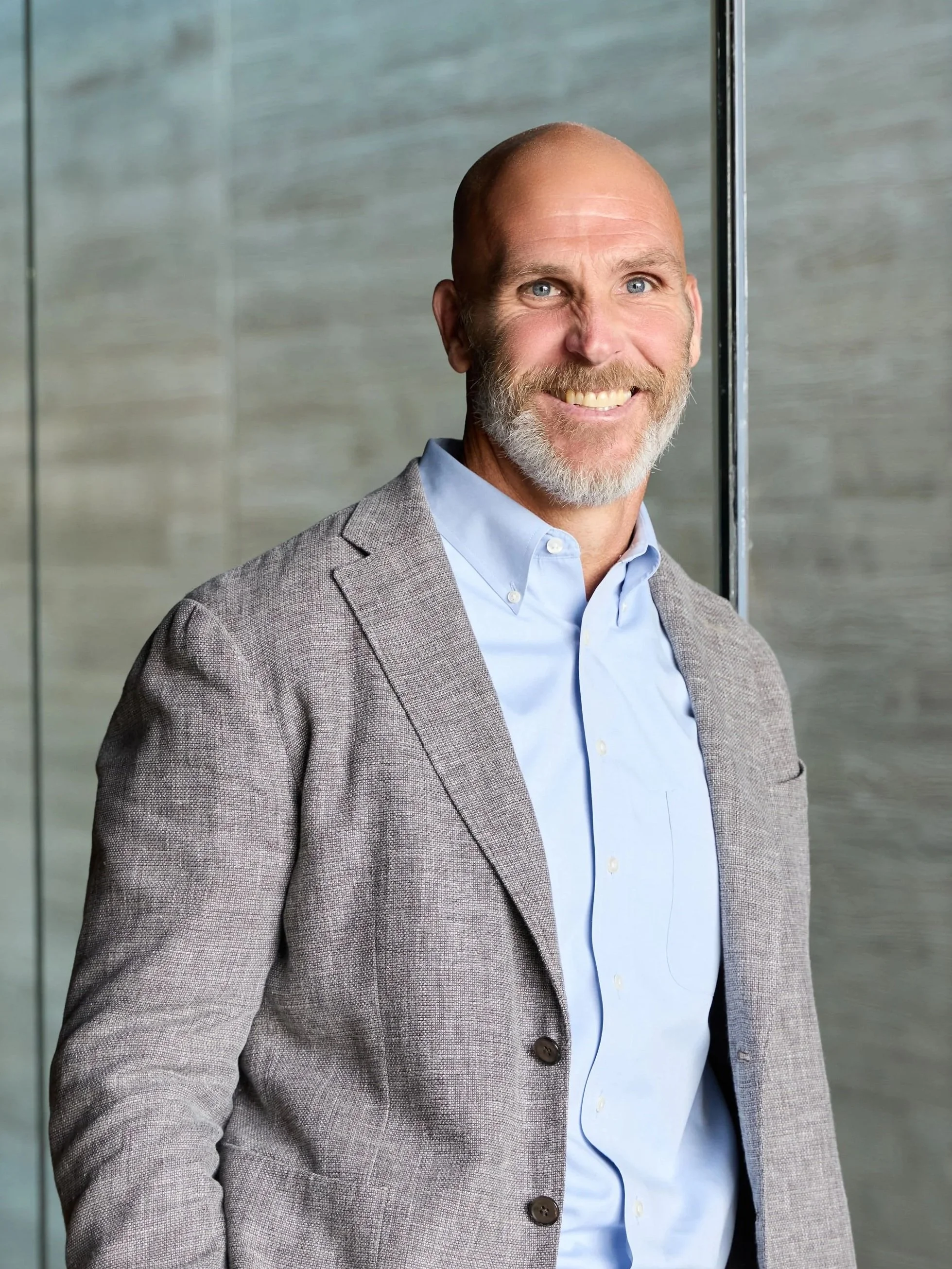 A smiling middle-aged man with a beard and short hair, wearing a light blue button-up shirt and a gray blazer, standing against a gray modern wall.