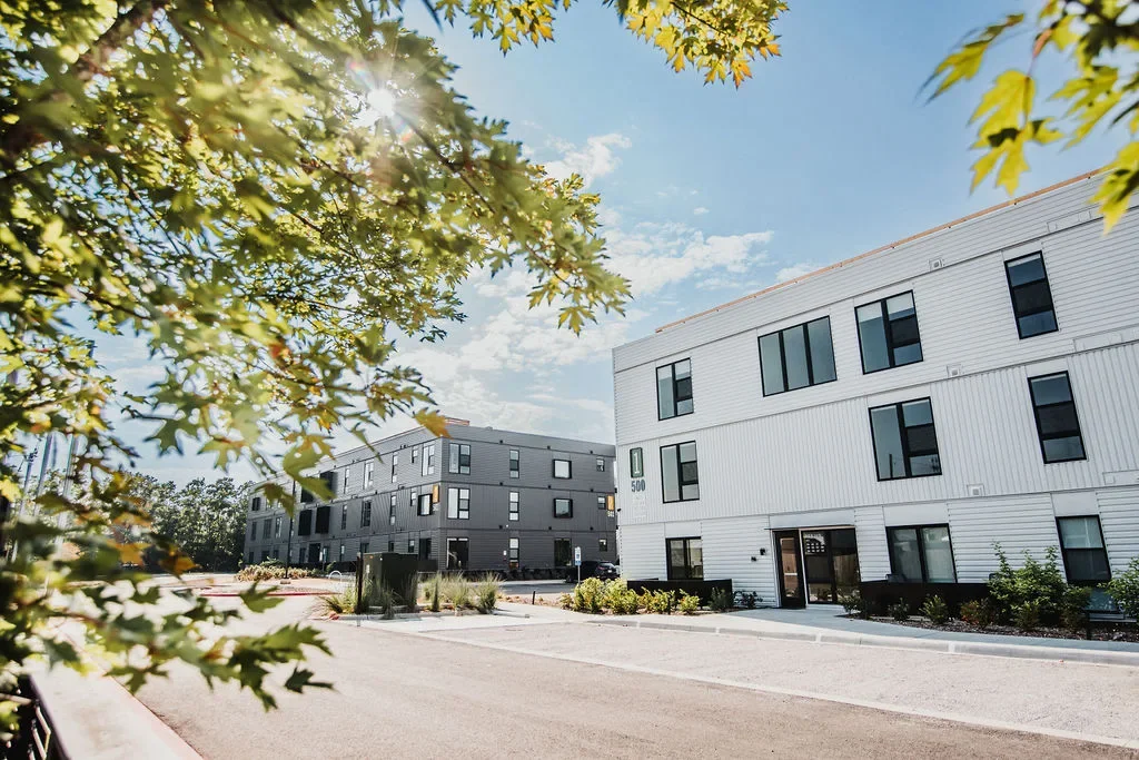 Modern apartment complex with white and gray exterior, surrounded by trees and landscaping, under a bright blue sky with scattered clouds.