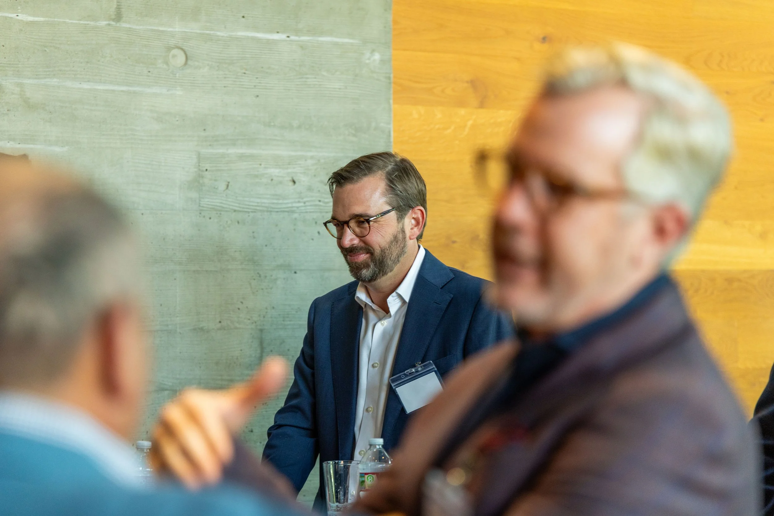 A professional man with glasses, a beard, and a suit is sitting at a table, smiling. Other people are in the foreground, blurred, in a discussion. The background features a split-colored wall with green and yellow wood panels.