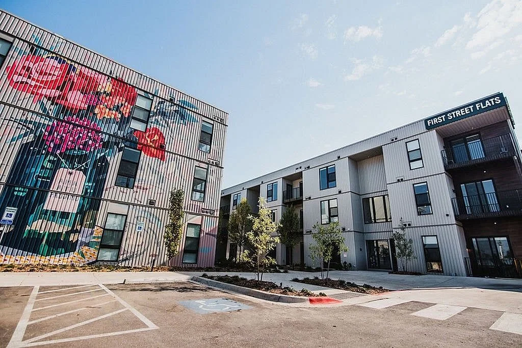 Modern apartment complex with a vibrant mural on the exterior wall and a sign that reads 'First Street Flats' near the entrance, with parking spaces and young trees in the foreground.