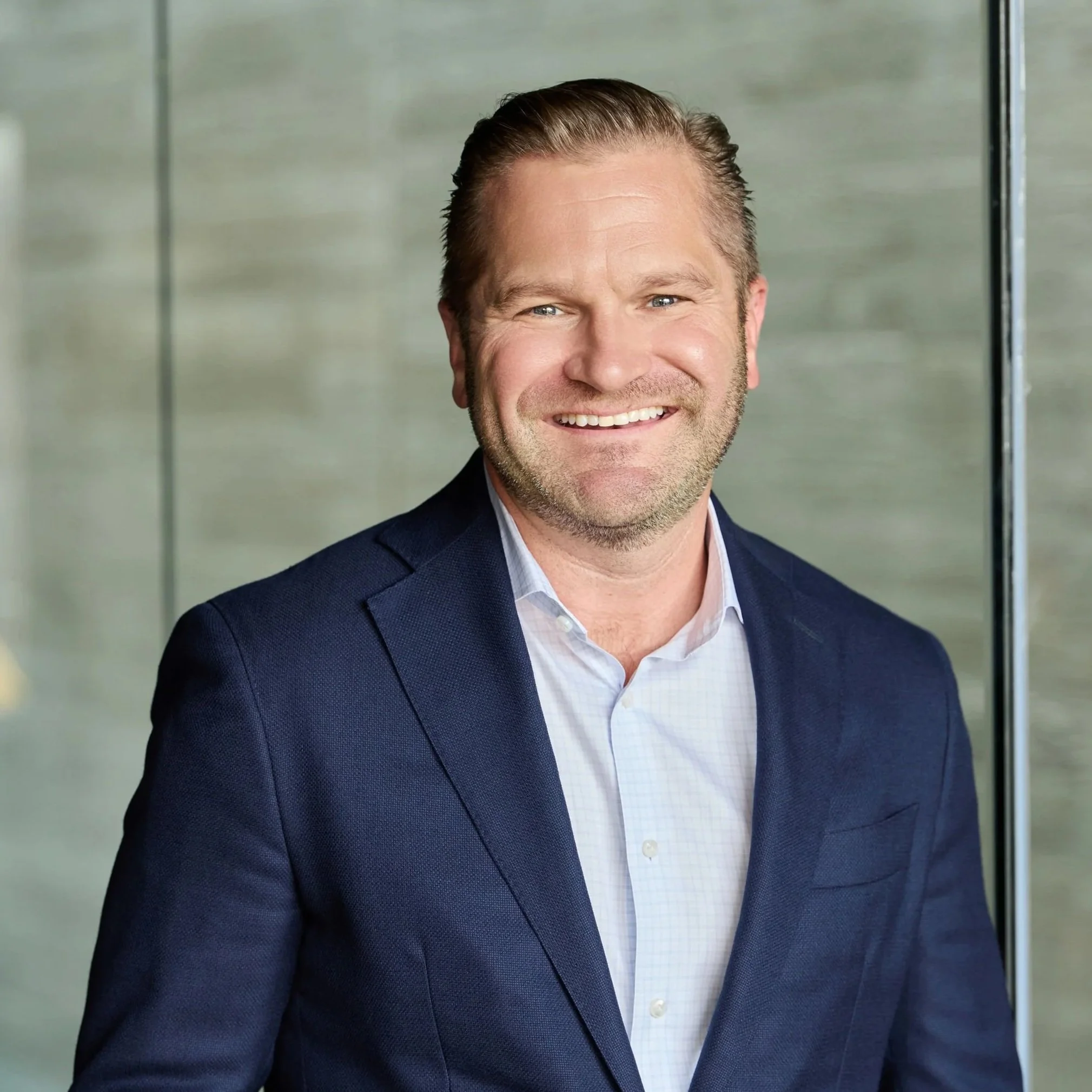 Portrait of a smiling man with short light brown hair, wearing a dark blue suit and a light blue collared shirt, standing in front of a modern blurred background.