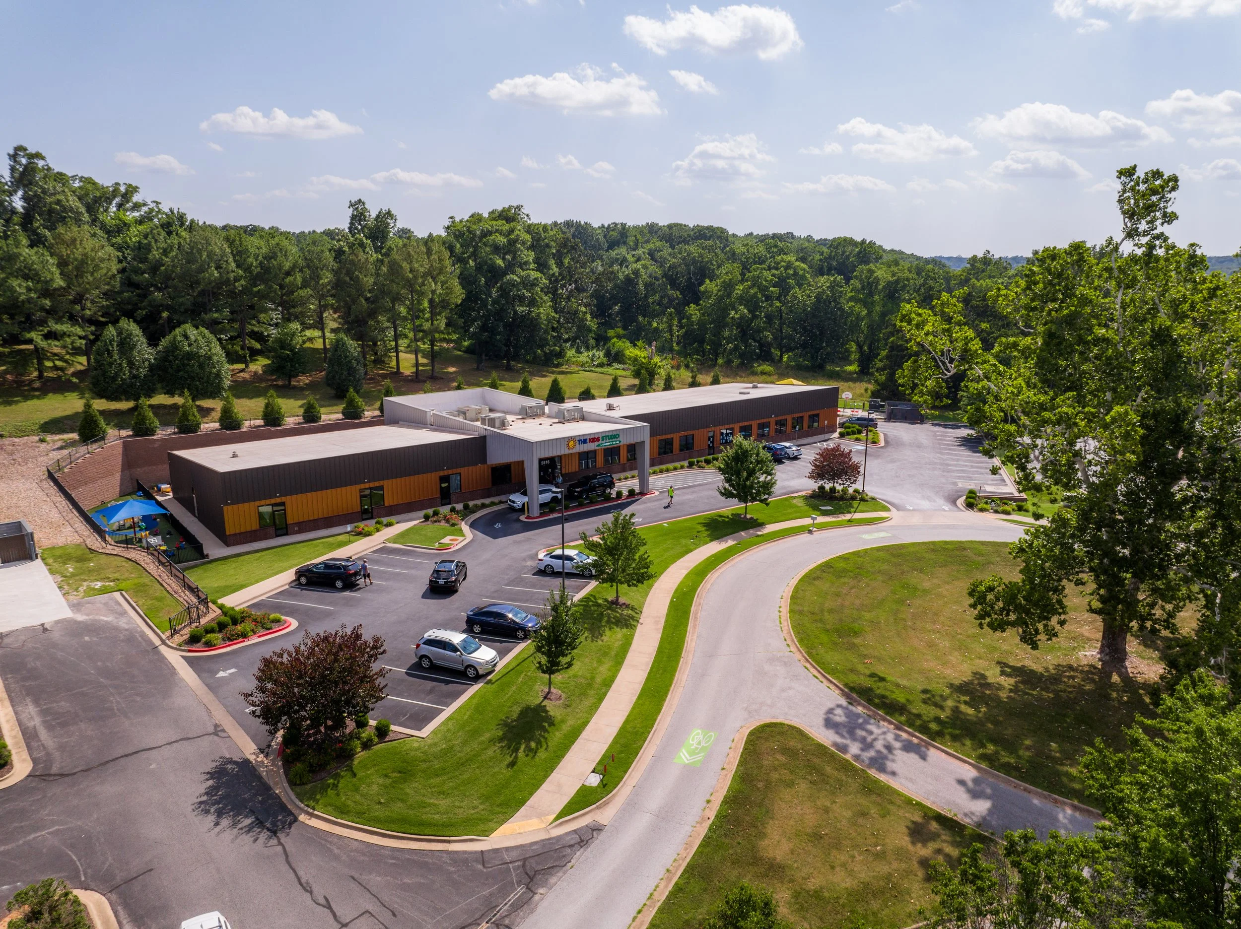 Aerial view of a single-story building with a parking lot, trees, and a surrounding green landscape under a partly cloudy sky.