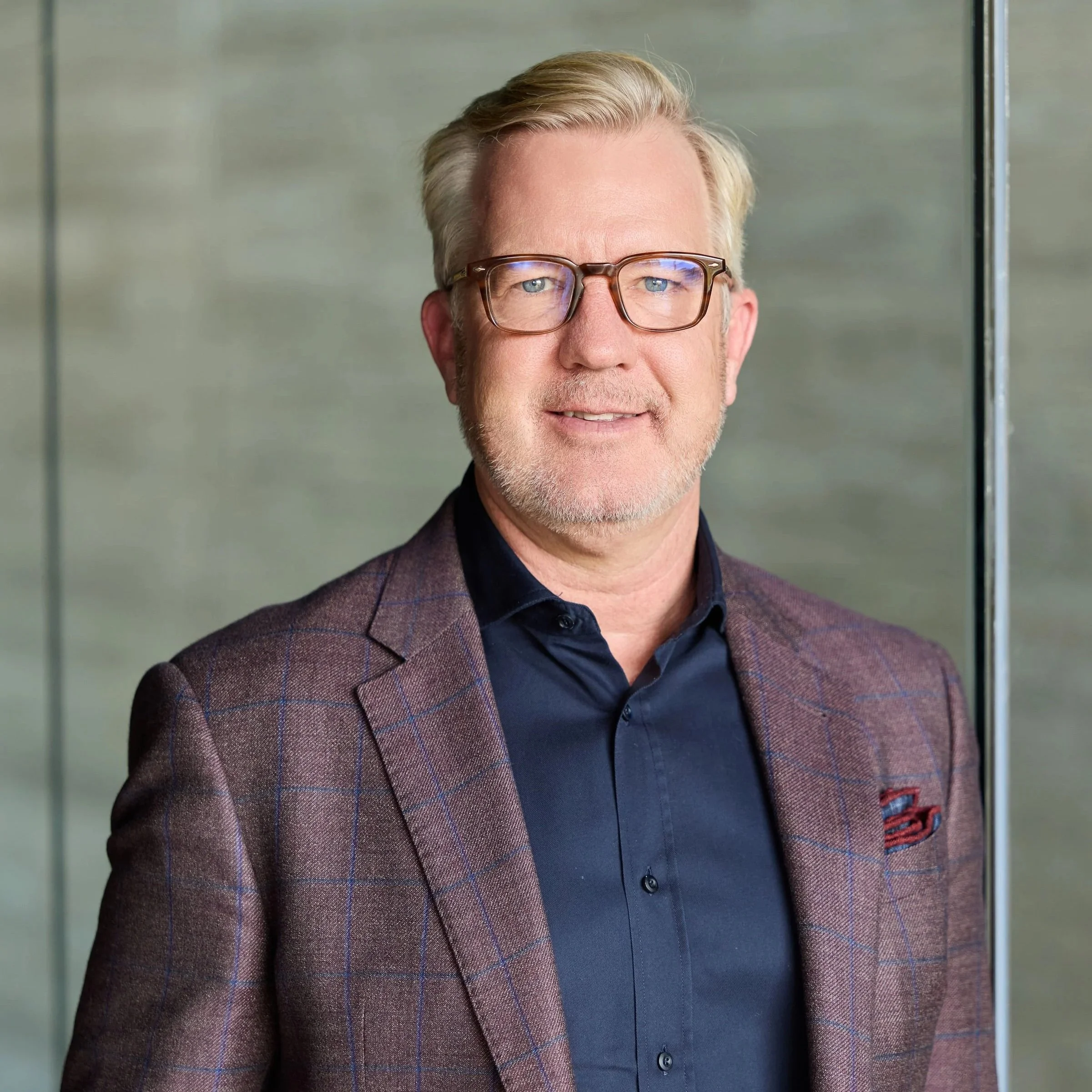 A middle-aged man in a checkered brown blazer and navy shirt, wearing glasses, standing indoors with a neutral background.