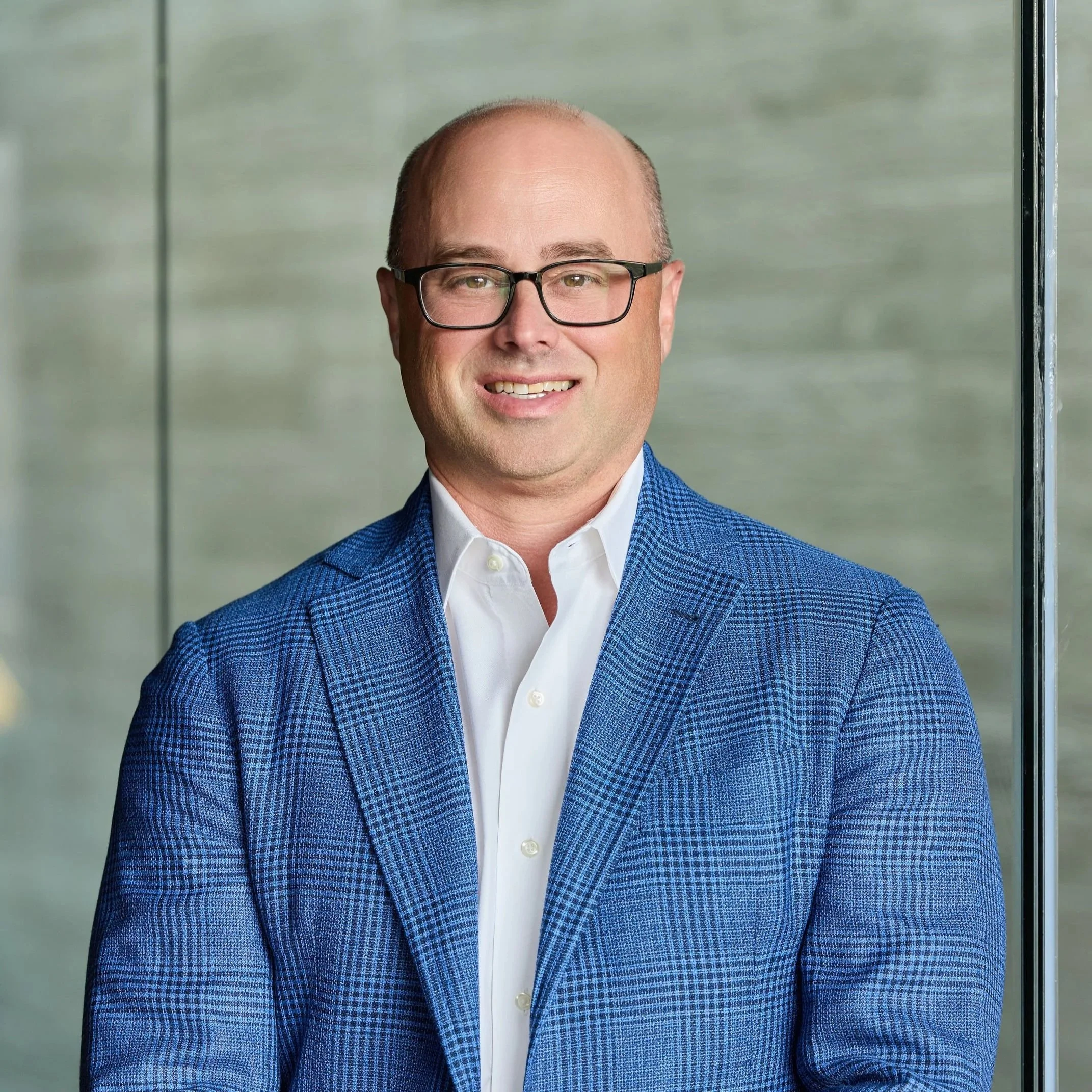A man wearing glasses, a white dress shirt, and a blue checked blazer, standing indoors near a large window with blurred background.