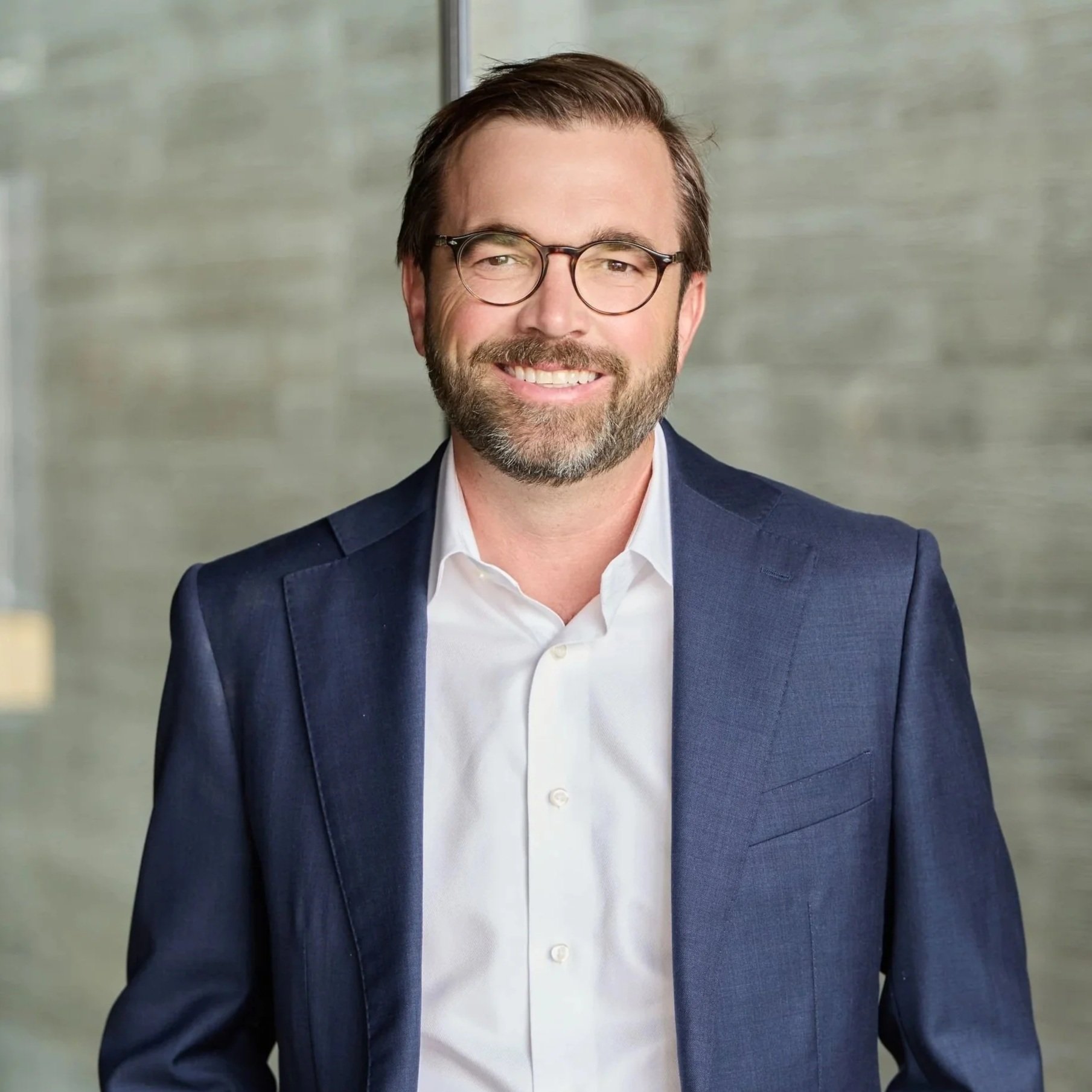 A man with glasses, a beard, and dark hair, smiling, wearing a navy blue blazer and white shirt, standing in front of a blurred indoor background.
