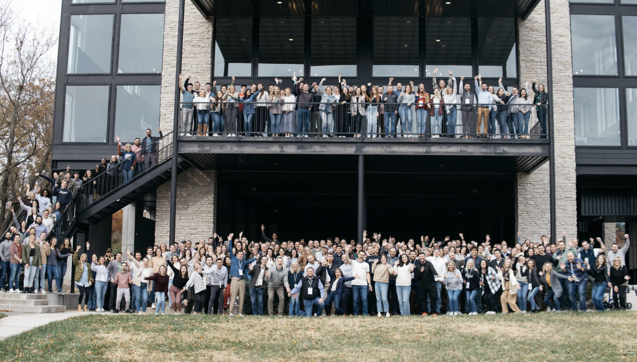 Large group of diverse people gathered outside a modern building, posing for a group photo on the lawn and balcony, with many waving or raising their hands.