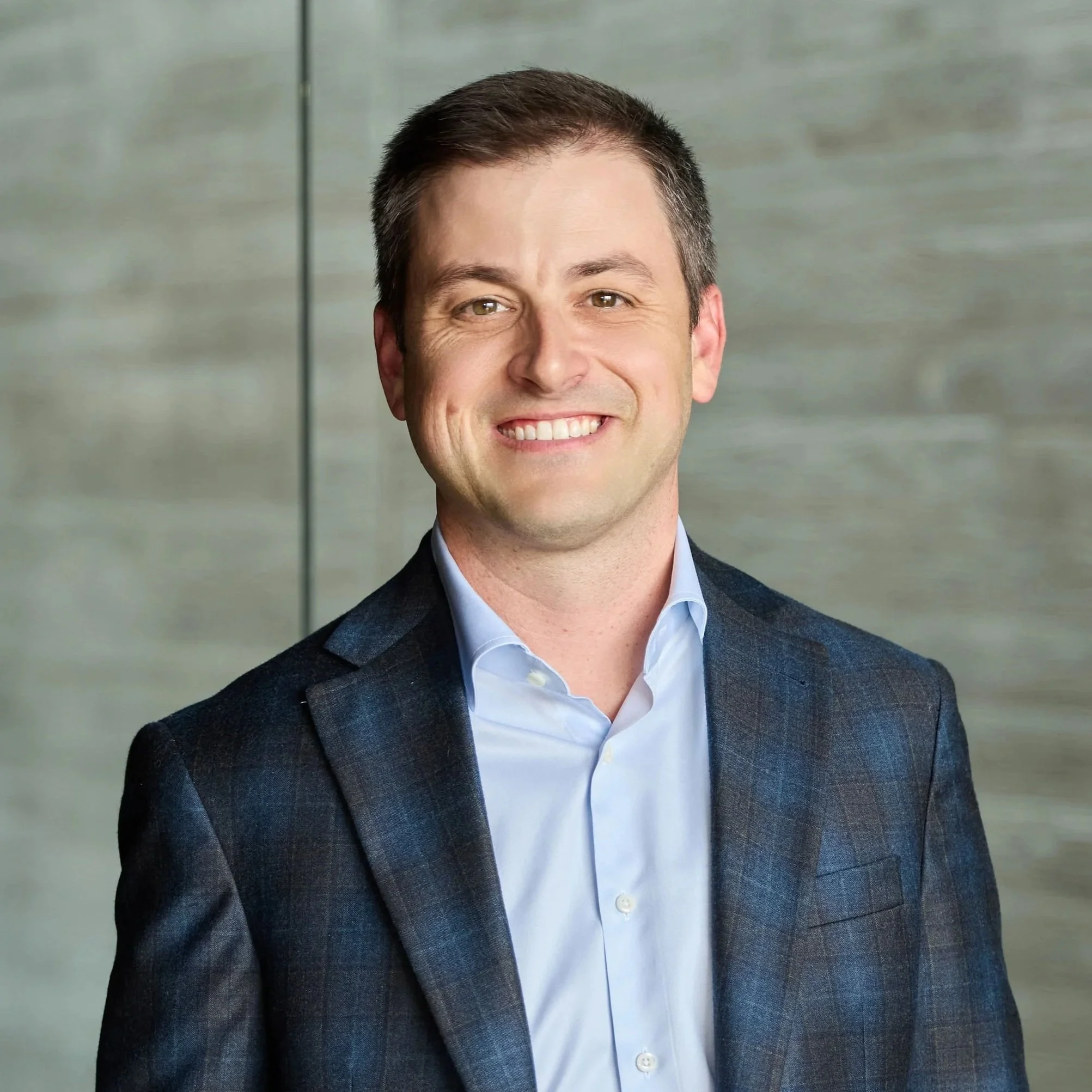 Headshot of a smiling man in a dark blazer and light blue shirt, standing indoors with a blurred background.