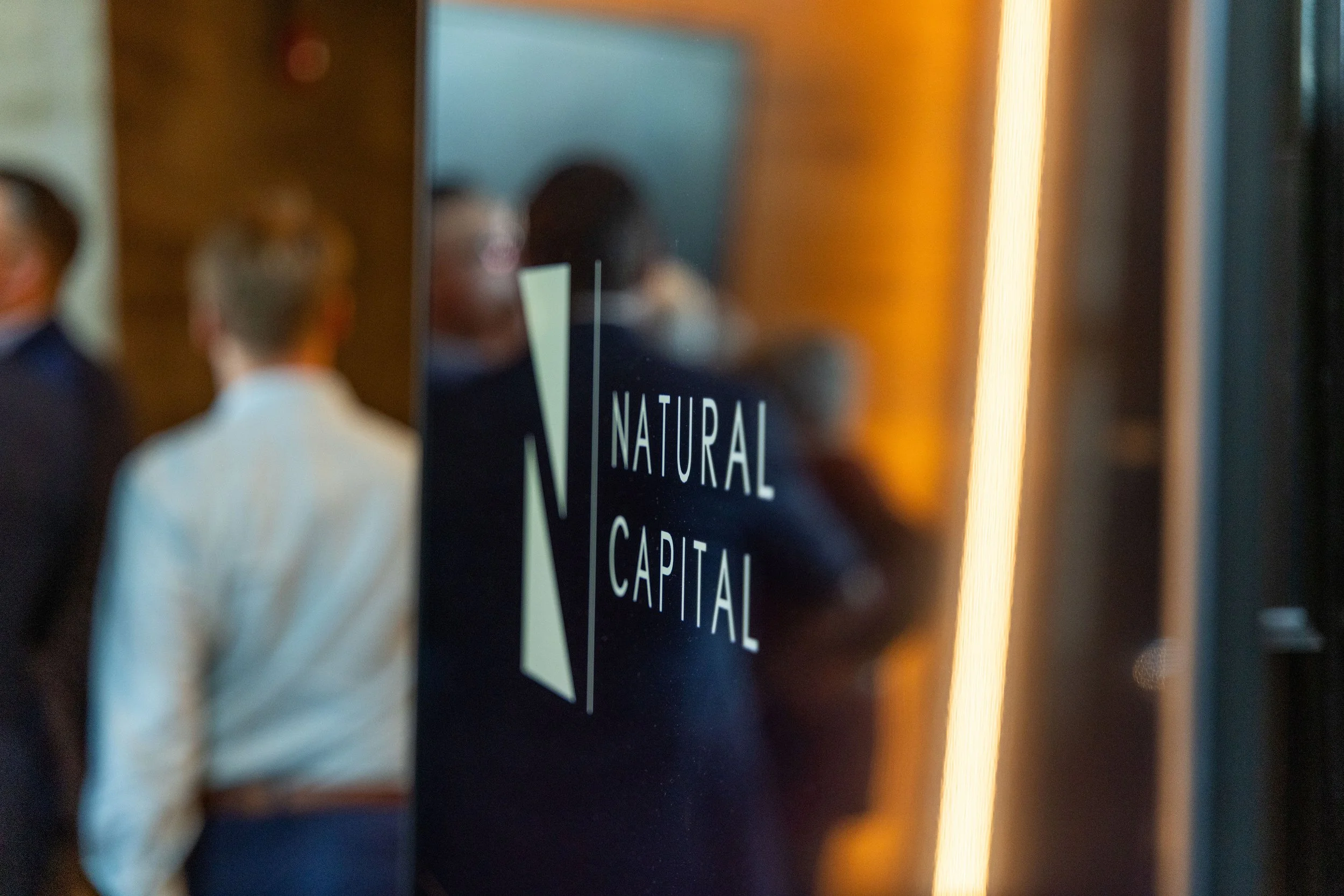 People in business attire reflected in a glass door with the words 'Natural Capital' on it.