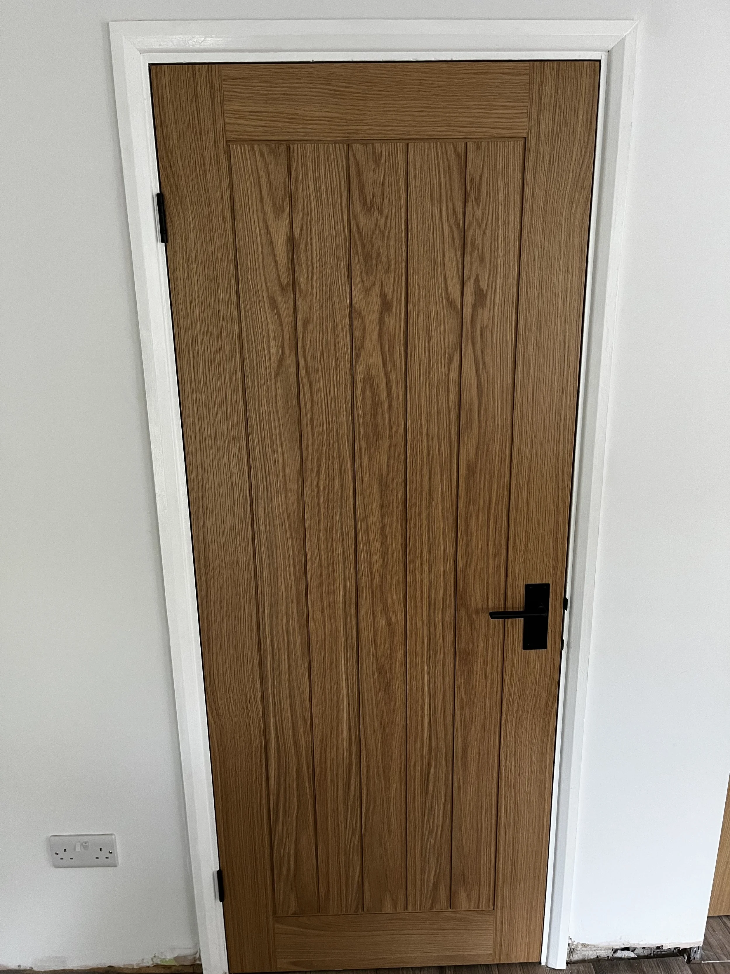 A wooden interior door with a black handle, framed in white trim, set in a white wall with electrical outlets nearby.