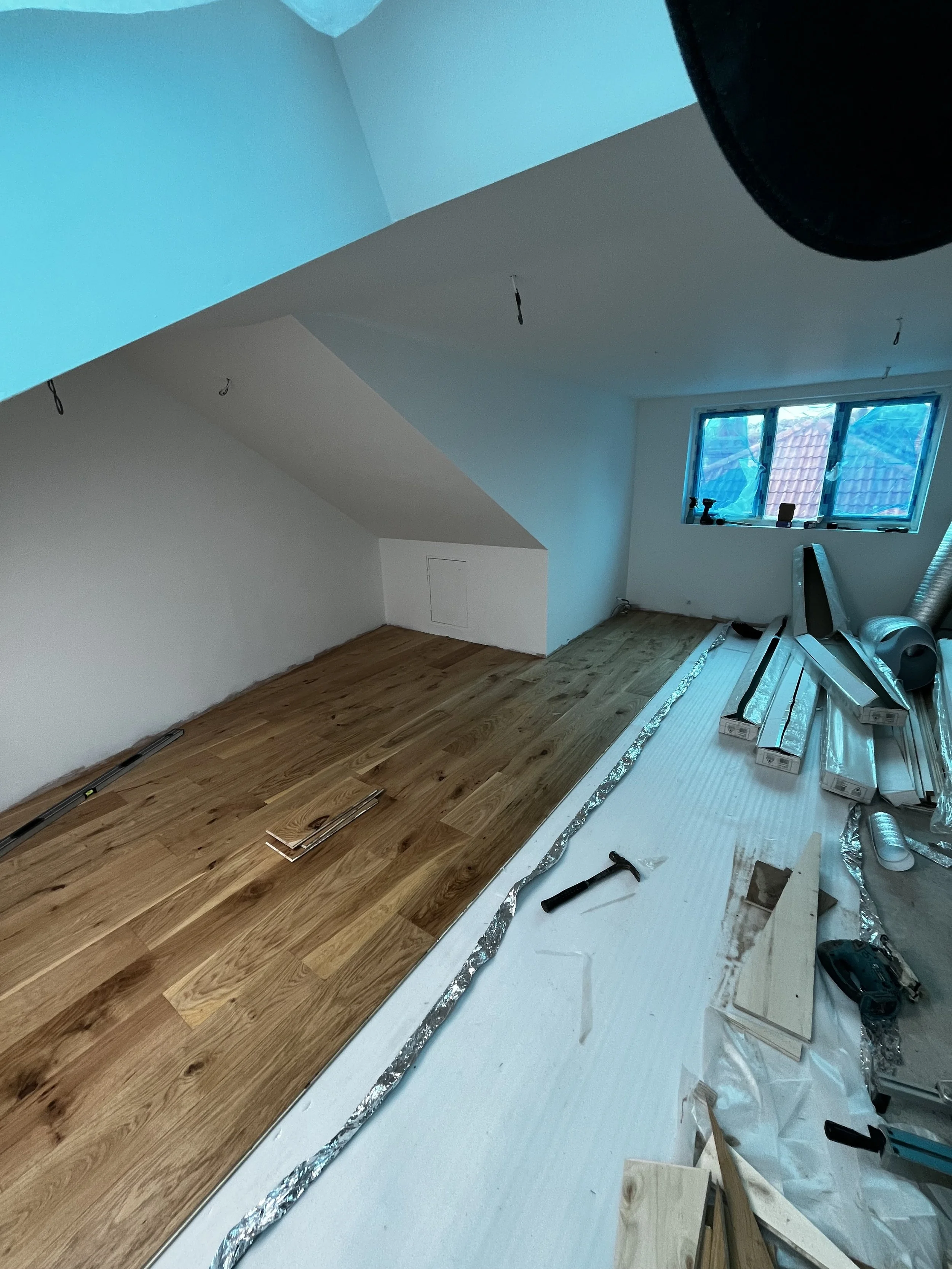 Attic room under renovation with wooden flooring partially installed, tools and supplies scattered around, and a window showing rooftops outside.