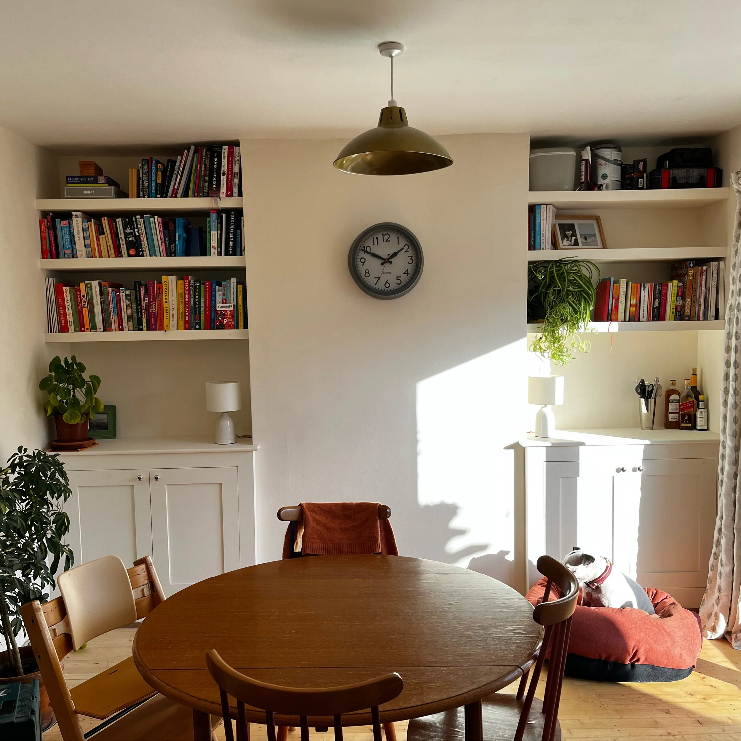 Dining room with a round wooden table, chairs, a white cabinet with plants and lamps, a wall clock, and bookshelves with books and decor, sunlight streaming in.