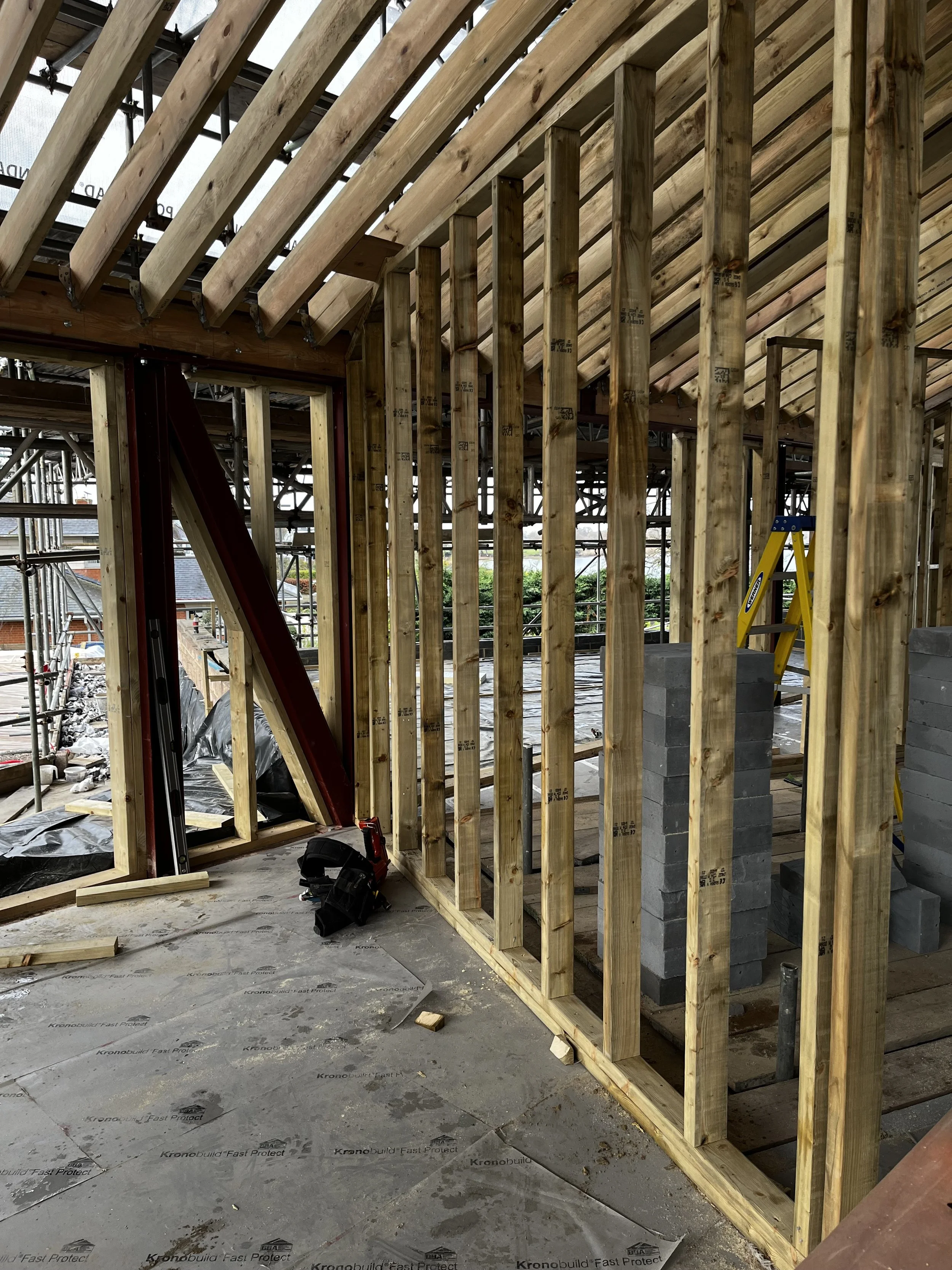 Interior view of a house under construction with wooden framing, a stack of concrete blocks, a yellow step ladder, and construction tools.