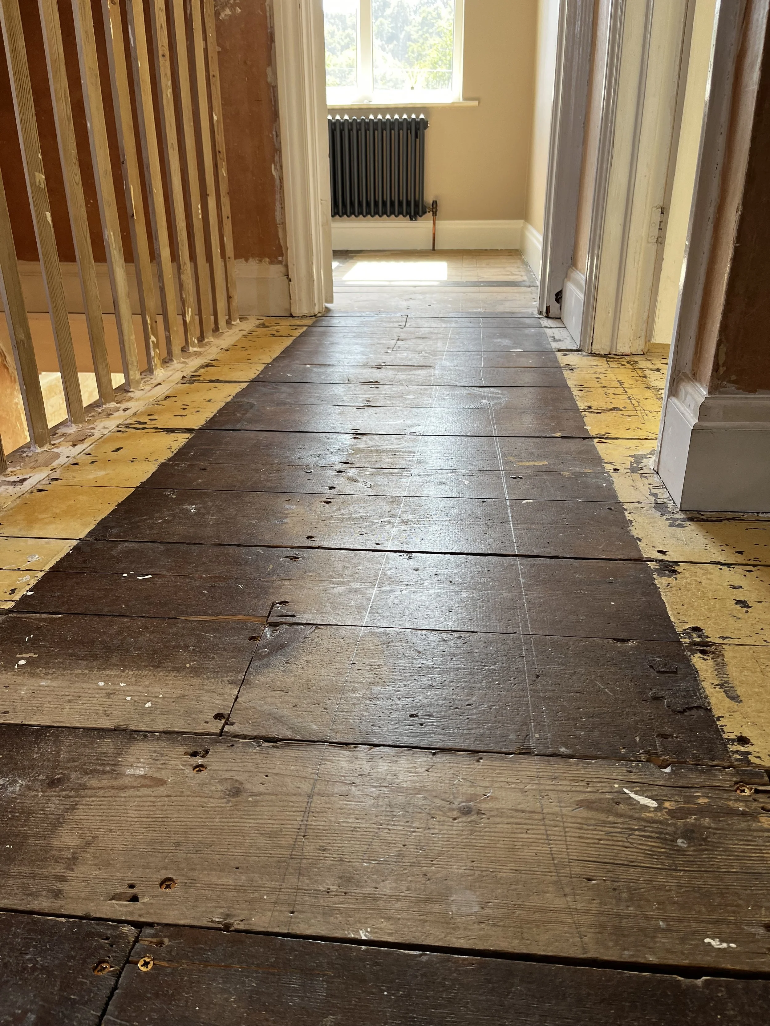 View of a house hallway under renovation, with unfinished wooden floorboards and visible wall studs on the left, leading to a sunlit room with a window and radiator.
