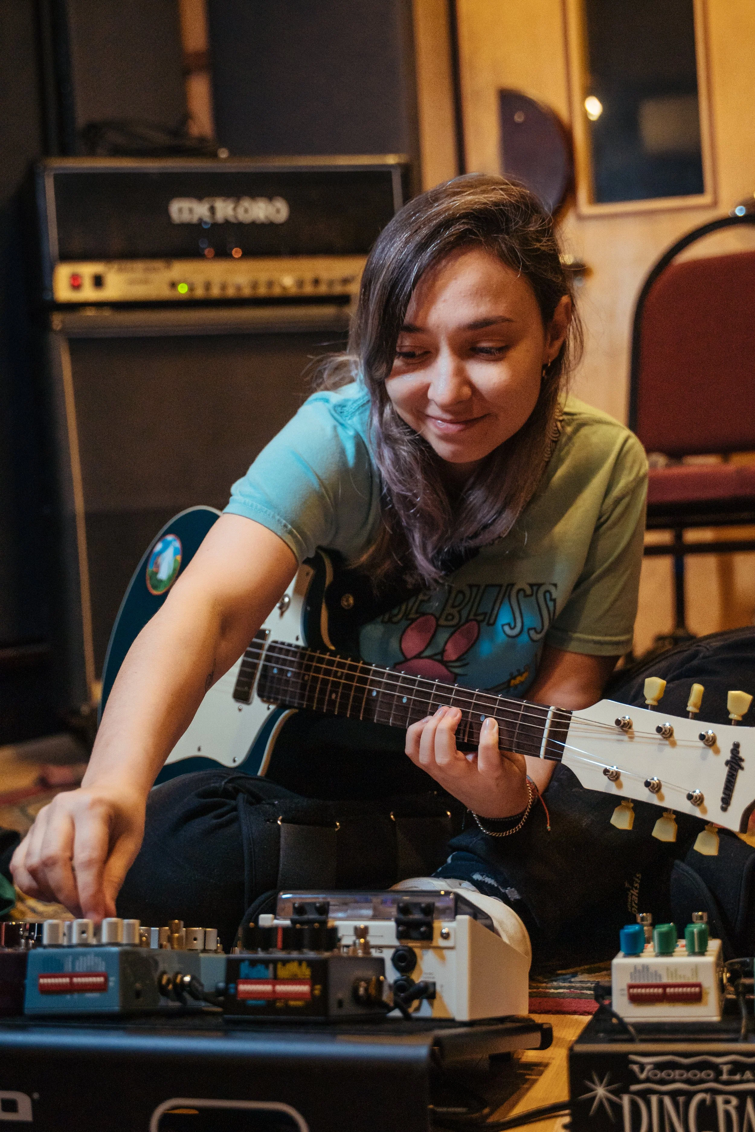 AJ playing guitar in a studio and adjusting effects on a Blooper pedal.  AJ tocando guitarra em um estúdio e ajustando efeitos em um pedal Blooper.
