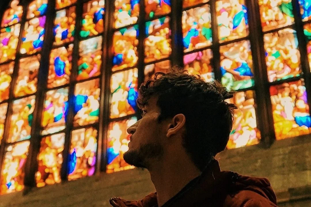 Un bărbat cu părul creț și barbă stă în fața unui vitraliu mare și colorat, în interiorul unei clădiri din Milano, Italia. A man with curly hair and beard standing in front of a large, colourful stained glass window inside a building in Milan, Italy.